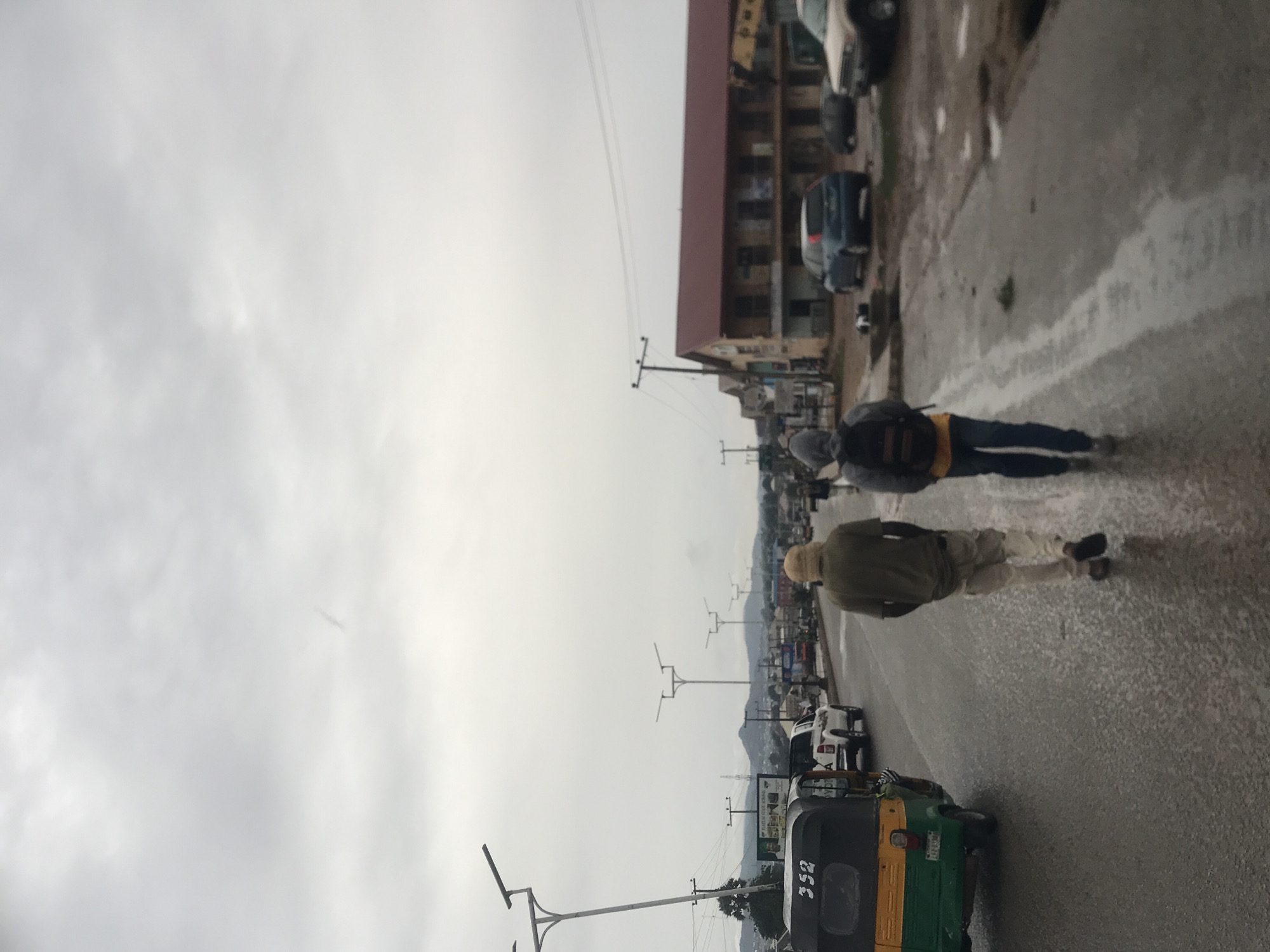 This image depicts a street scene in Rafin Par, Nigeria, under a uniformly overcast and grey sky. The road surface appears wet with puddles and reflections, indicating recent rain.
In the foreground, on the left side of the road, is a green and yellow auto-rickshaw (often called a 'Keke Napep' in Nigeria) with the number "352" printed in white on its rear. Two individuals are walking away from the camera on the right side of the wet asphalt. The person closer to the camera wears a light-colored head covering, an olive green top, and light trousers. Further down the road, another individual wears a dark hooded jacket, a backpack, and dark trousers, with a yellow shirt visible underneath.
The street is lined with utility poles carrying overhead wires, and modern-looking streetlights with multiple arms extend over the road. Along the right side of the street, various buildings are visible, some multi-story with reddish-brown roofs. Several cars are parked or driving along the road in the mid-ground and background. In the distance, a range of hazy hills or mountains is discernible against the cloudy horizon. A blurred sign on the right side of the road displays partial text, which appears to include "MADU... FOTE GLOBAL". The scene captures a typical daytime urban or semi-urban setting, likely after a downpour.