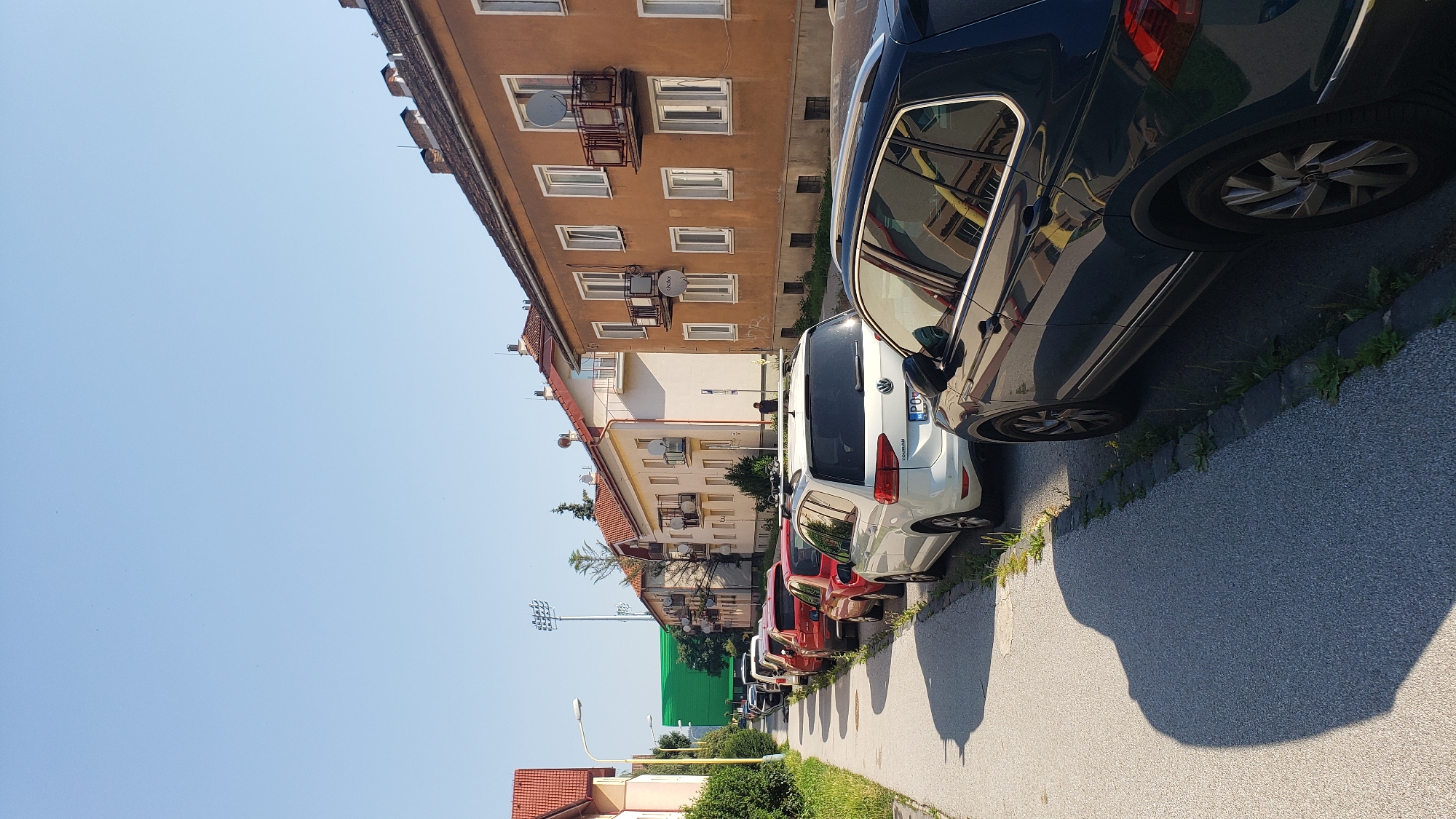 A street-level, eye-level, medium shot captures a row of parked cars lining a residential street on a bright, sunny day. The cars are parked parallel to the sidewalk on the right side of the frame, with the closest car being a dark blue SUV. Behind it, a white Volkswagen Touareg is parked, followed by several other cars of various colors, including red, white, and black. The street is paved, and a sidewalk runs along the left side of the image, bordered by grass and some small weeds. In the background, a row of apartment buildings with orange and white facades are visible. These buildings have multiple windows and balconies. To the far left, a building with a red roof and white walls is partially visible. Further in the distance, a tall green structure, possibly a screen or a large advertisement, stands out against the clear blue sky. Adjacent to the green structure are stadium lights on tall poles. The overall scene depicts a typical residential area in a city. This is a photograph of Šarišská ulica in Prešov, Slovakia.