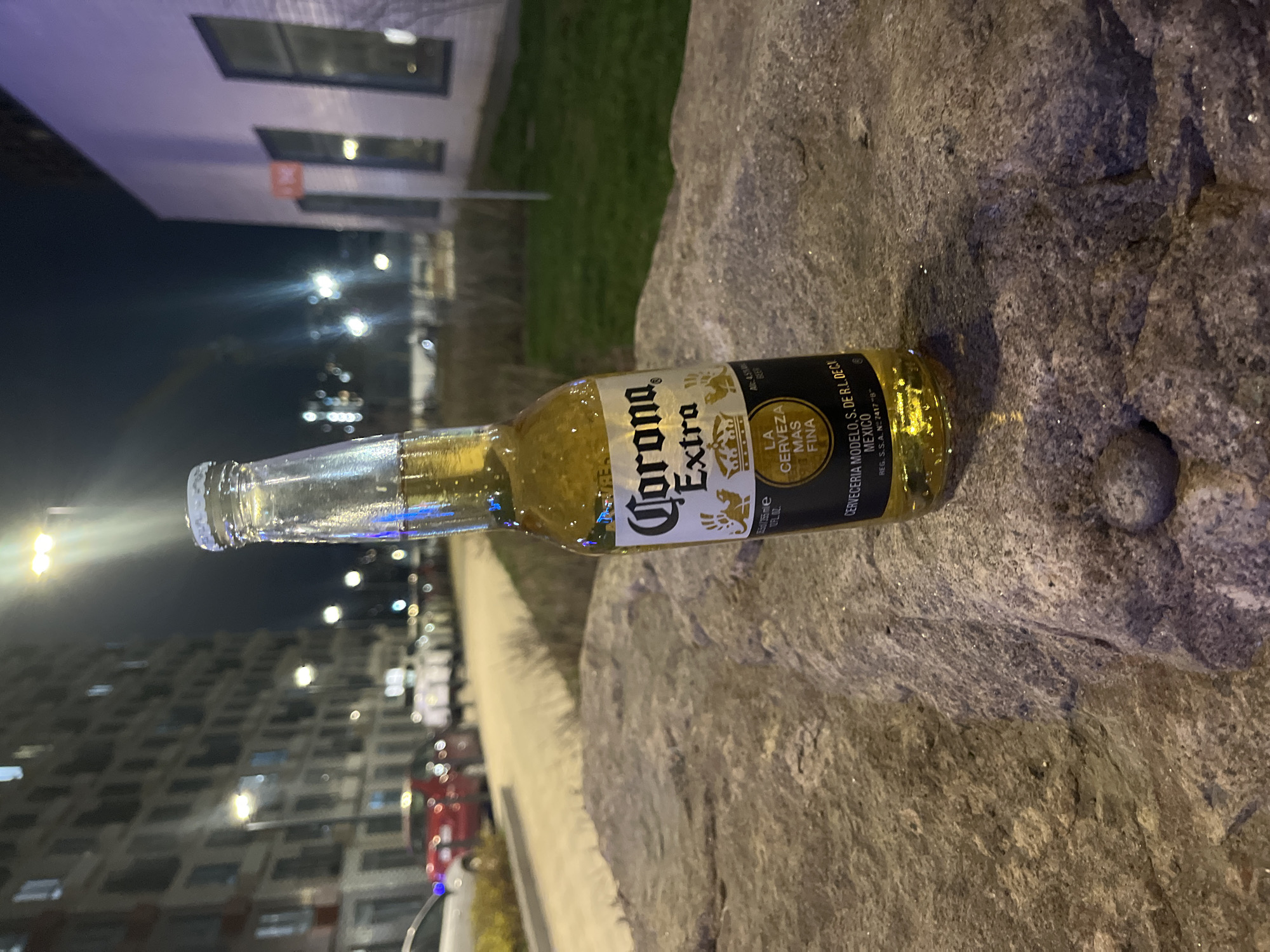 The image features a Corona Extra beer bottle lying horizontally on a coarse, grey-brown stone surface. The bottle is partially filled with a golden liquid and contains visible bubbles. Its label clearly displays "Corona Extra," along with the phrases "LA CERVEZA MAS FINA" (The Finest Beer), "CERVECERIA MODELO, S. DE R.L. DE C.V.," and "MEXICO." Other details visible on the label include "REG. S.S.A. N: 7417 "B"," "12 FL. OZ.," "Alc. 4.5%," and "BEER."
The scene is set at night or late evening, illuminated by various artificial light sources. In the background, modern urban architecture is visible, with multi-story buildings featuring numerous windows. One building to the upper left has a light-colored facade with dark, rectangular windows, and a sliver of green grass can be seen nearby. Further in the distance, other residential buildings and a street or parking area are discernible. Bright, streaky light sources, likely streetlights or distant car lights, create an urban glow against the dark sky. Partial views of parked cars, including red and white vehicles, can be observed.
No people are present, and no specific activity is depicted, presenting a quiet, contemplative urban moment. The overall setting points to an urban environment, specifically located in Kudrovo, Russia.
