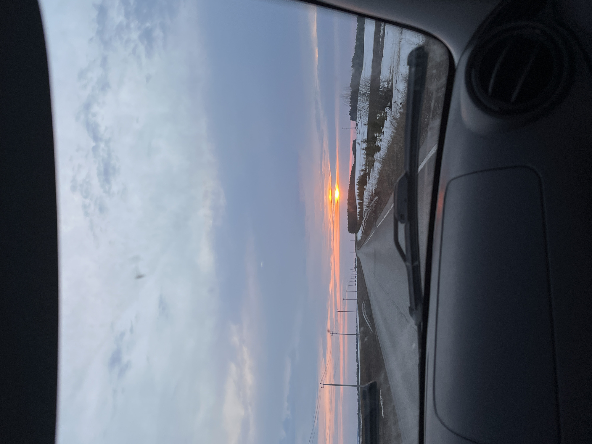 The image is taken from inside a vehicle, looking out the front windshield. It is daytime. The sun is setting, visible down a long, straight road. The road is flanked by what appears to be snowy or icy fields. Utility poles run parallel to the road on the left side. The interior of the vehicle is visible on the right side of the frame, including part of the dashboard and an air vent. The location is Ledukhi, Russia, based on the provided information.