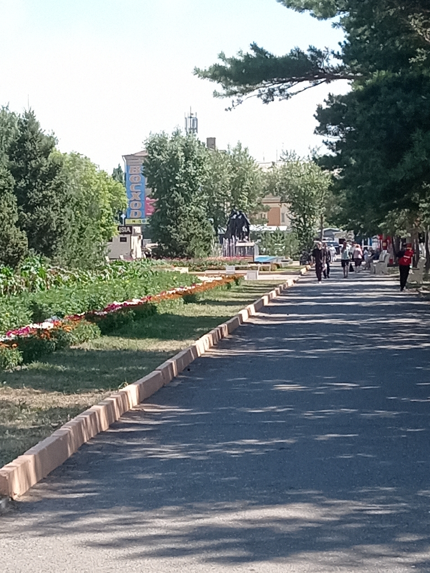 The image depicts a paved pathway lined with trees and a flowerbed in Rudny, Kazakhstan. Several people are walking on the pathway and sitting on benches. In the background, a building with a large blue and yellow sign is visible, along with a statue of what appears to be horses. The sky is clear and blue, and the overall scene suggests a public park or a pedestrian area on a sunny day.