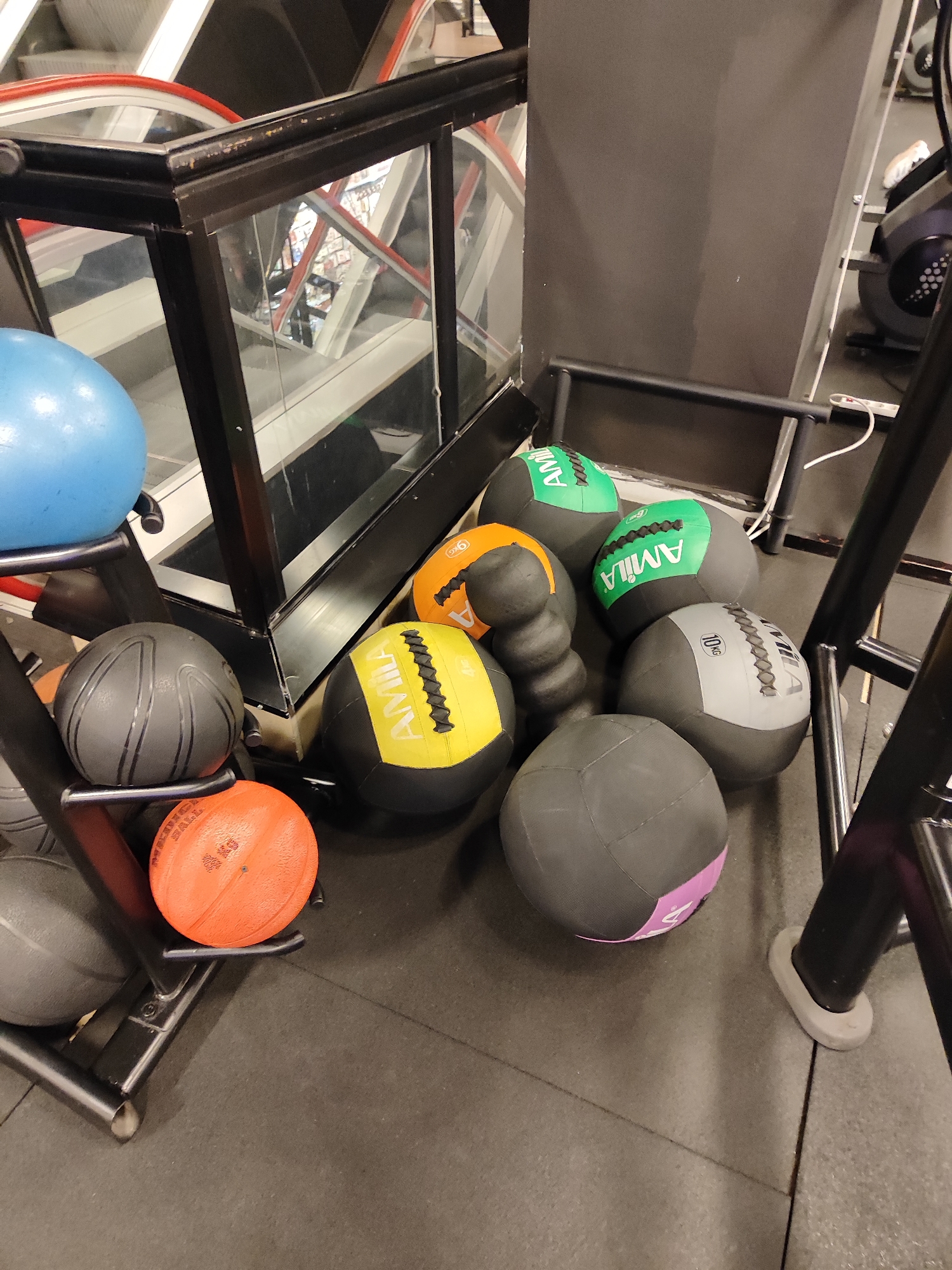 The image displays a collection of exercise balls and gym equipment in an indoor fitness facility, likely located in Belgrade, Serbia. In the foreground, a black metal rack holds several balls: a large, smooth, bright blue exercise ball, a dark gray medicine ball with prominent ridged stripes, and an orange medicine ball clearly labeled "4 KG" and partially "MEDICINE BALL."
On the dark rubber gym floor, a variety of other medicine and slam balls are scattered. These include multiple gray and green balls branded "AMILA" with "8 KG" visible, a yellow and black "AMILA" ball, an orange and black ball marked "9 KG," a gray and white "AMILA" ball displaying "10 KG," and a gray and purple ball. A stack of small, ribbed, black training accessories is also present among the balls on the floor.
In the background, a structure of black metal framing and glass panels reflects a red handrail, suggesting an escalator or moving walkway is nearby. To the right, a white power strip with a cord and a part of a black exercise machine are visible. The setting is illuminated by artificial light, and no people are present, implying the equipment is either stored or ready for use.
