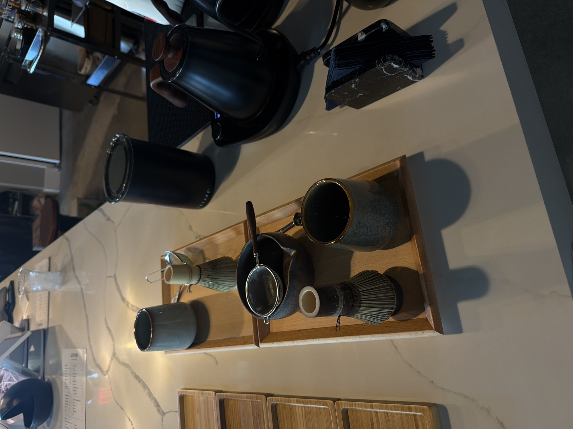 This image depicts a countertop display of tea-making accessories, likely in a cafe or home setting, located in North Miami Beach, United States of America. The scene is dominated by a marble-patterned countertop, reflecting ambient light.
On the left side of the frame, a stack of wooden trays is visible. In the center, a wooden tray holds a collection of matcha tea preparation items. These include a blue ceramic cup, a bamboo whisk (chasen), and a small sieve with a wooden handle resting over a dark ceramic bowl. Further down the tray are another bamboo whisk and a dark green ceramic cup.
To the right of the tray, a black electric kettle with a wooden handle sits on a black charging base. Adjacent to the kettle is a black cylindrical container, possibly for tea leaves or storage. In the foreground on the right, a black marble-patterned napkin holder with dark blue napkins is positioned on the countertop. The background is blurred, showing glimpses of shelves with various items and a stool. The overall impression is a neatly arranged setup for preparing and serving matcha tea.