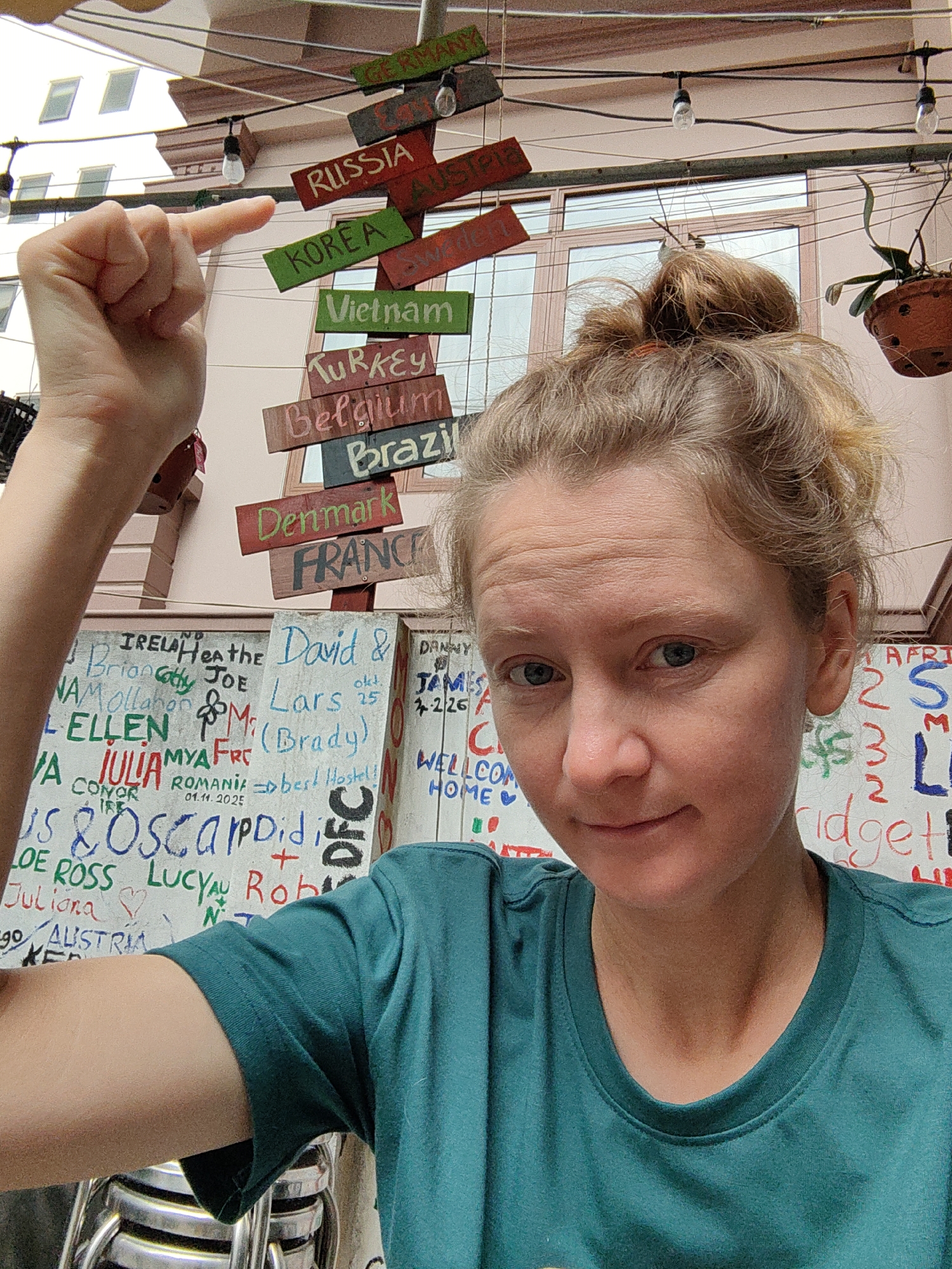 A woman with blonde hair in a messy bun and wearing a teal t-shirt is taking a selfie in Bai Chay Ward, Vietnam. She is pointing her finger towards a signpost with country names, including Germany, Russia, Korea, and Vietnam. The background includes a wall with handwritten messages and illustrations, string lights, and a hanging potted plant.