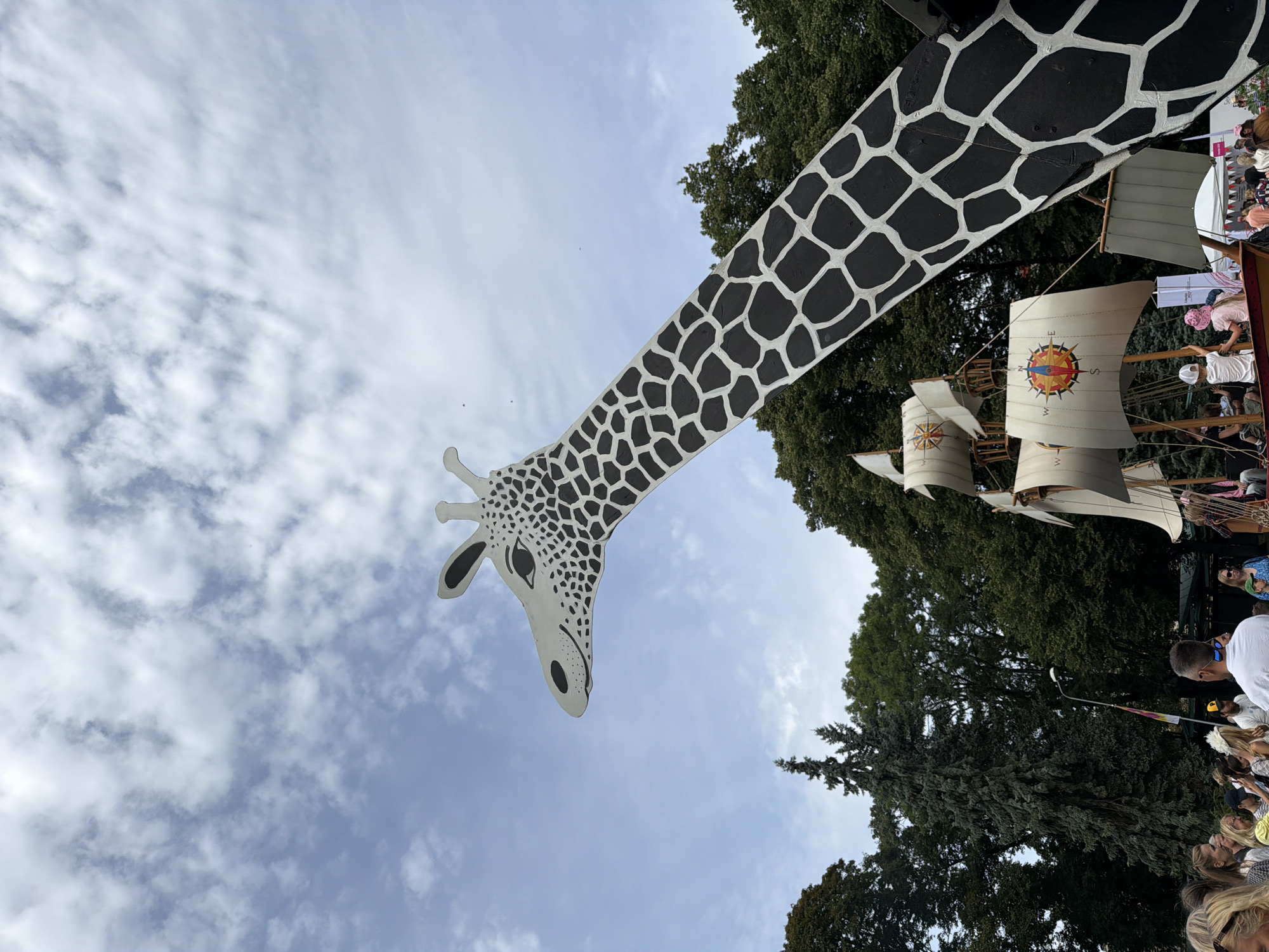 The image depicts an outdoor event, possibly a festival or exhibition, in Riga, Latvia. A prominent feature is a large, stylized sculpture of a giraffe's head and neck, painted with black spots on a white background, extending into the sky. Below the giraffe, several boat-like structures with sails are visible, some adorned with compass rose symbols. A crowd of people, dressed in casual summer attire, are gathered around, suggesting an event open to the public. The background consists of lush green trees and a cloudy sky. One of the boat sails has "LIDL" and "2021" printed on it.