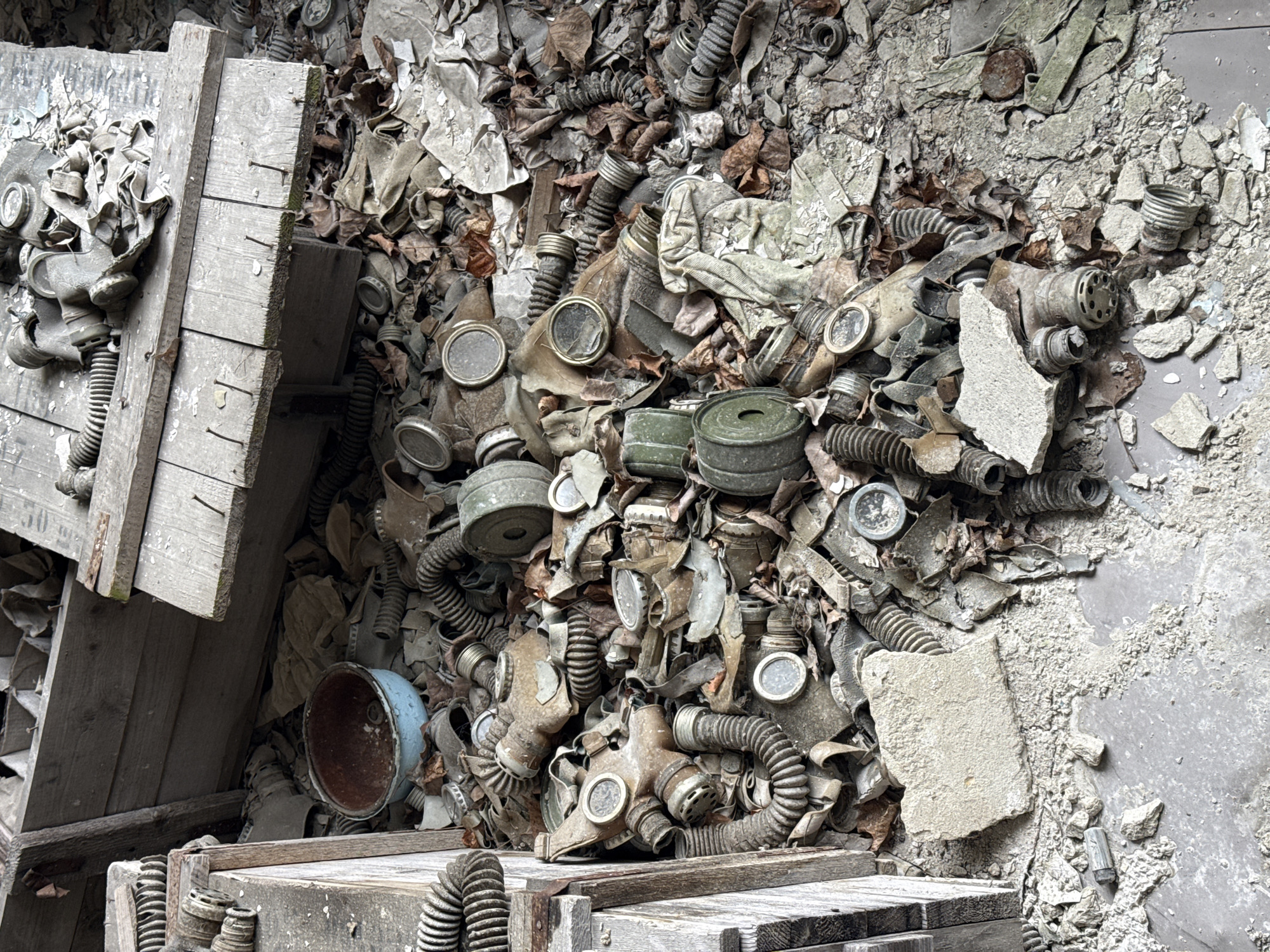 The image depicts a debris field of discarded gas masks and associated equipment inside a building in Prypiat, Ukraine. A partially collapsed wooden structure is visible to the left. The masks are of varying models, showing signs of age, corrosion, and disintegration. The environment includes scattered leaves, paper, and building debris, indicating abandonment and decay. There are no people visible in the scene.