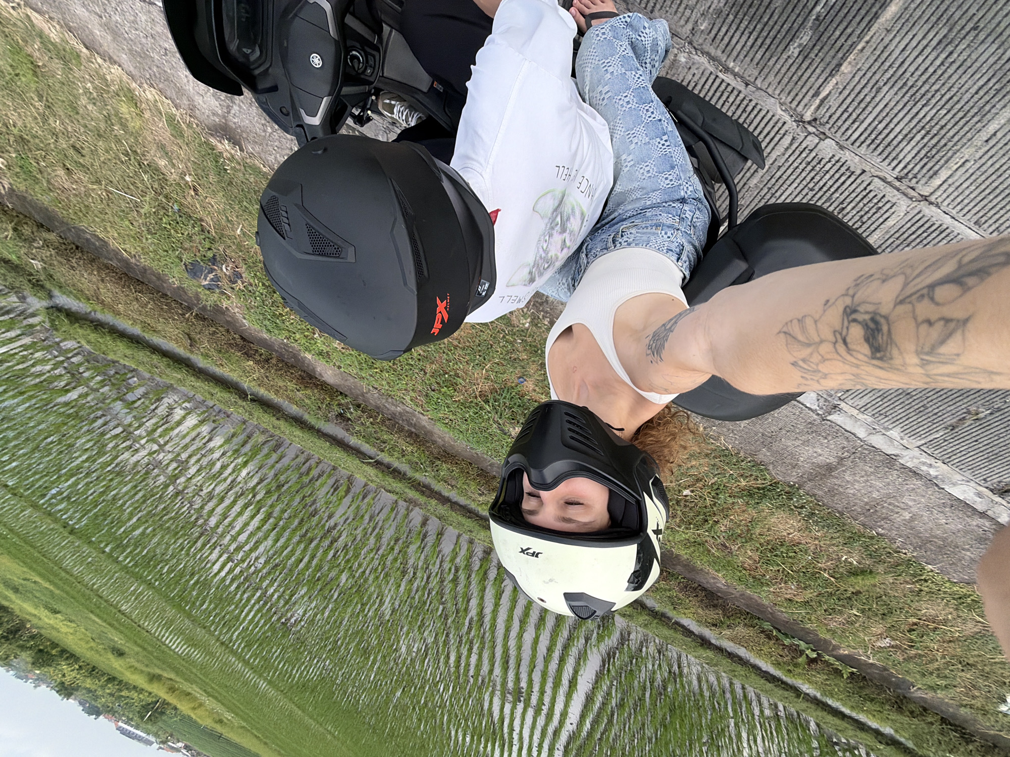 Two people, wearing helmets, are seated on a motorcycle. The person in front is wearing a white tank top and patterned blue jeans, and has curly brown hair. The person behind is wearing a white long-sleeved shirt and black pants. The motorcycle is parked on a paved surface next to a lush green rice paddy. Buildings and trees are visible in the distance. The scene appears to be in North Kuta, Indonesia, a region known for its rice fields.