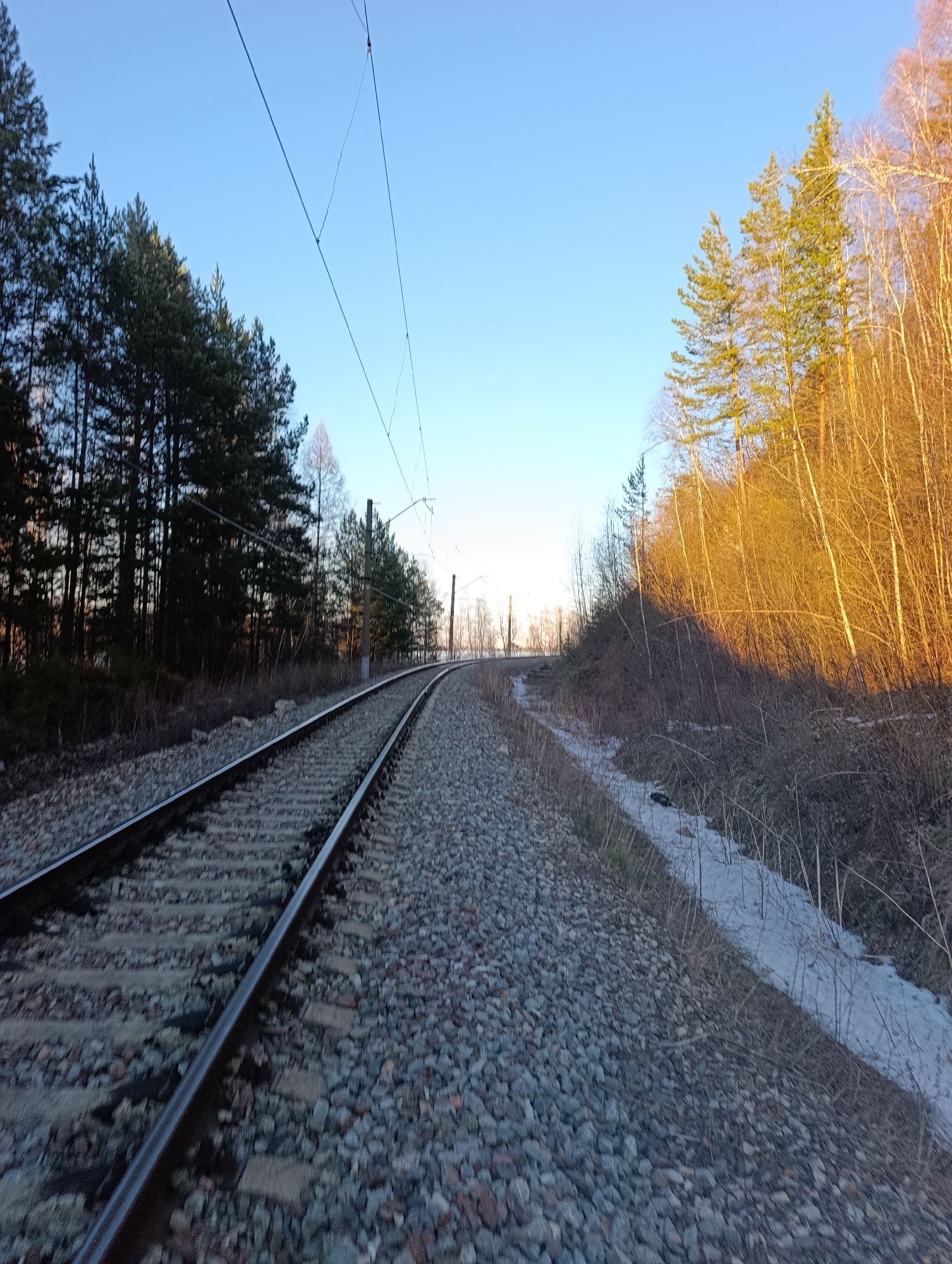 The image depicts a railway track curving into the distance in a wooded area. The track is laid on a bed of gray gravel. Overhead electrical lines are visible, running parallel to the tracks. To the left of the tracks, a dense forest of coniferous trees stands. To the right, the terrain slopes downward with a mix of deciduous trees and lingering patches of snow. The sky is clear blue, indicating daylight. Based on visual cues and possible location metadata, this could be a section of the railway system on the outskirts of Krasnoyarsk, Russia, passing through a forested area. No people are visible in the frame.