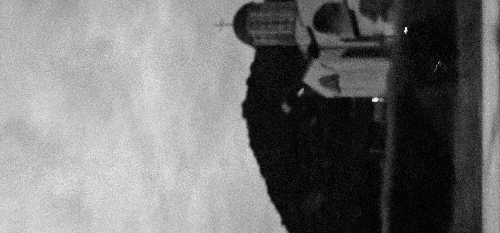 This is a grainy, high-contrast black and white photograph featuring an architectural structure. The image appears to have been taken in monochrome, or converted to it, and exhibits significant visual noise, giving it a rough, textured appearance.
The primary subject is a large, dark, imposing structure occupying the right side of the frame. It appears to be made of stone or concrete, with visible details suggesting architectural elements like columns or patterned surfaces, although clarity is limited by the grain. A distinct cross symbol is visible at the very top of the structure, indicating it could be a church, a monument, or another religious building.
The left and upper-left portions of the image consist of a uniform, textured light grey area, likely representing an overcast sky or a brightly lit, undifferentiated background. The lighting is soft and diffuse, with no harsh shadows, suggesting either an overcast day or a bright ambient light source. There are no discernible people or specific activities taking place. The overall mood is stark and possibly somber due to the monochrome palette and the nature of the subject. No visible text is present in the image.