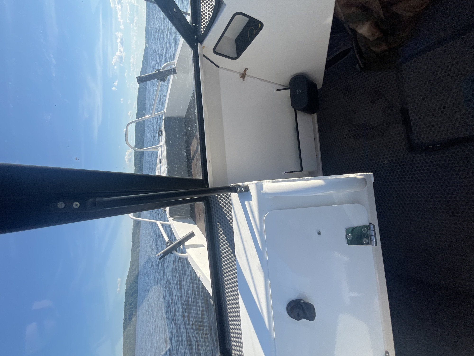 The image shows the interior of a boat navigating on a body of water, with land visible in the distance. The boat is white with black trim and has a windshield. In the foreground, there is a white storage compartment with a black latch and a metal hinge. The floor of the boat is covered with a black, textured material. A black object with the word "PUSH" is visible on the right side of the boat. The water is calm with gentle ripples, and the sky is blue with scattered white clouds. The land in the distance appears to be covered in green trees. Based on the context, the boat is likely located on the river near Siukterka, Russia, a village situated on the banks of the Tom River in the Kemerovo Oblast.