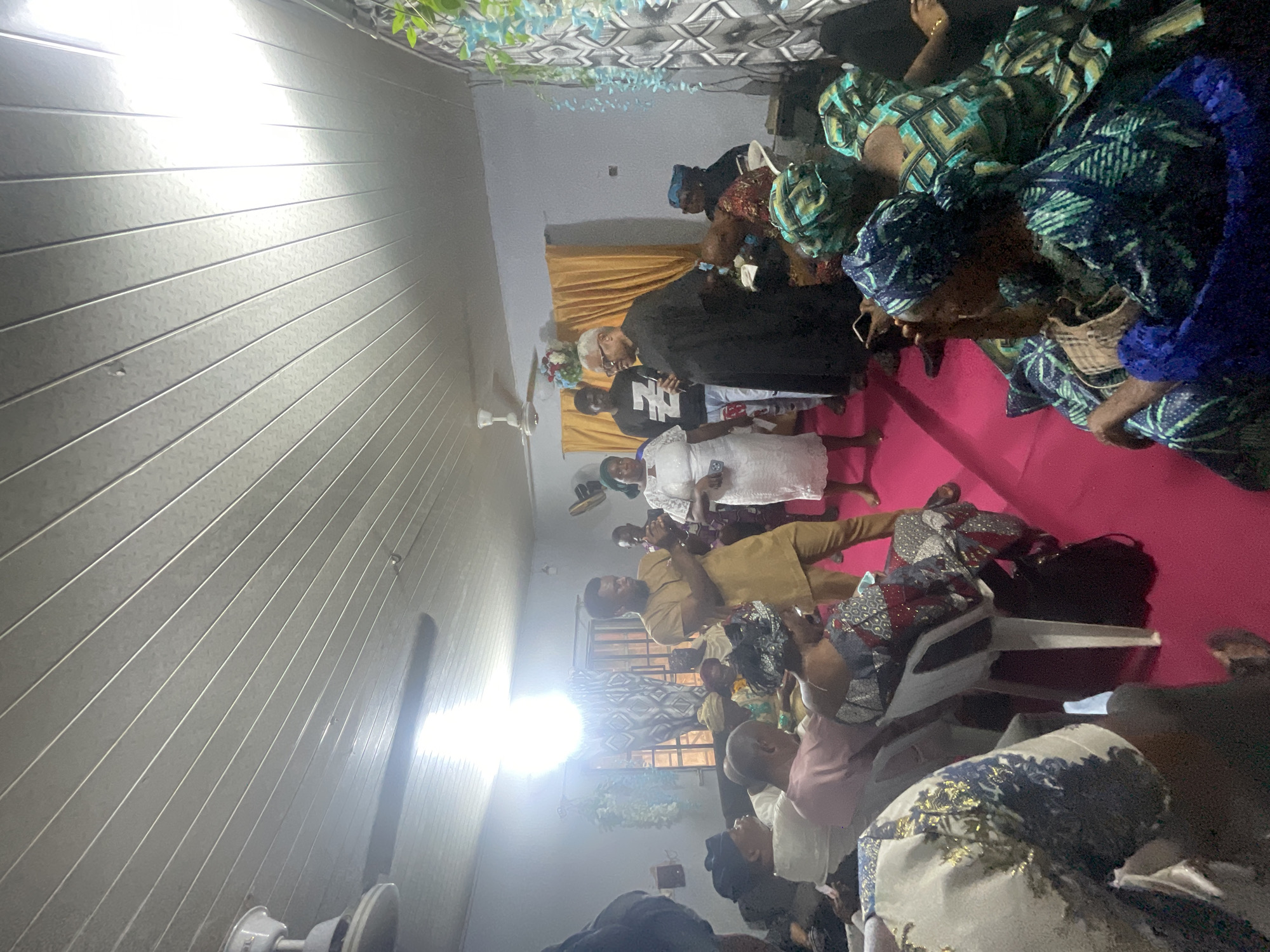 This image captures an indoor gathering featuring numerous people, predominantly adults, in what appears to be a hall or event space. The room's walls are covered with light-colored, horizontally paneled material, likely PVC, exhibiting a textured pattern. A prominent, bright artificial light source on the left side of the frame creates significant glare and illuminates parts of the room, with another light source visible in the background. A white ceiling fan is partially visible in the upper left.
Many of the attendees are women dressed in colorful traditional Nigerian attire, including patterned Ankara fabrics and matching headwraps (gele). Men are also present, with one in a light brown or golden tunic and another in a black t-shirt that displays the visible letters "N" and "Z". A woman in a white lace dress stands near the center. The floor is covered with a vibrant red or pink carpet, and several white plastic chairs are occupied by guests, while others stand and observe. Floral decorations, composed of light blue/green foliage and small flowers, are draped along the back wall, positioned above a golden-yellow curtain that acts as a backdrop.
The people appear attentive, suggesting they are engaged in an activity such as listening to a speaker or participating in a ceremony. The scene implies a celebration, meeting, or religious service, consistent with an event taking place in Lokoja, Nigeria. The strong artificial lighting indicates an indoor setting, possibly in the evening or a space with limited natural light.