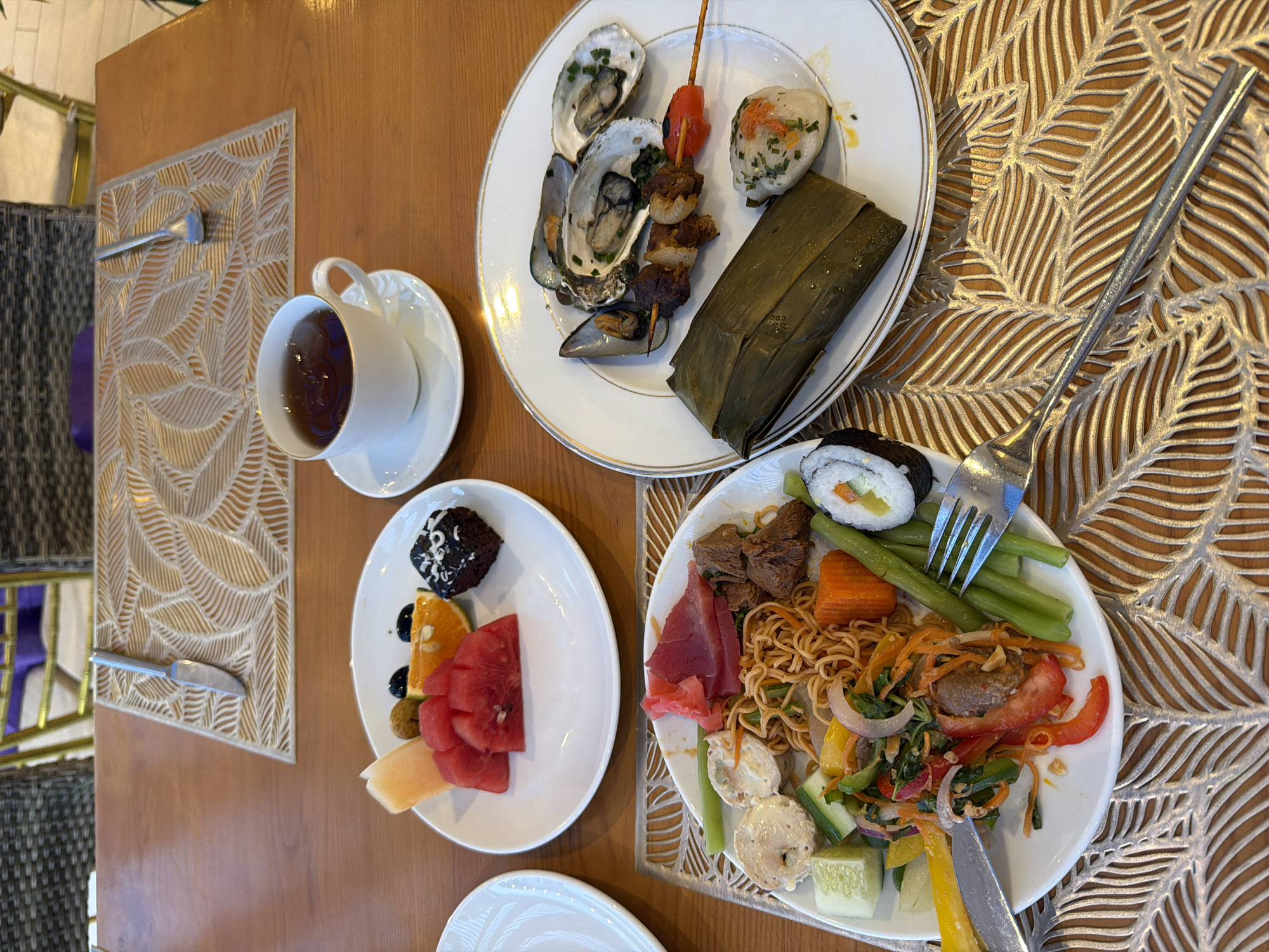 This image depicts a diverse meal laid out on a wooden table in Dong Ninh Hoa Ward, Vietnam. The scene is set on a light brown wooden table, featuring two woven placemats with a leaf pattern.
On the table, three white plates with gold rims are present.
The top-middle plate contains sliced watermelon, a piece of cantaloupe, an orange segment, two dark olives, a small unidentified light-colored fruit, and a square chocolate brownie topped with white icing.
To the left-bottom, a large plate holds an assortment of food including noodles, various mixed vegetables (such as cucumber cubes, bell pepper strips, and shredded carrots), pieces of cooked meat, sushi maki rolls, slices of raw tuna (sashimi style), pickled ginger, and several pale, round items (possibly dumplings or fish cakes). A fork and knife rest on this plate.
To the right-middle, another plate features two oysters on the half shell garnished with herbs, one mussel, a meat skewer (kebab) with meat and a red cherry tomato, a rectangular item wrapped in a banana leaf, and a small, round, white item topped with green chives.
A white teacup with a matching saucer, containing a dark beverage, is positioned between the top-middle and right-middle plates.
Cutlery includes a butter knife and a spoon placed on the top placemat. A large spoon is partially visible on the bottom-right placemat.
In the background, parts of woven chairs are visible, along with a golden-colored chair frame and a purple cushion, suggesting an indoor dining environment. No individuals are visible in the frame.