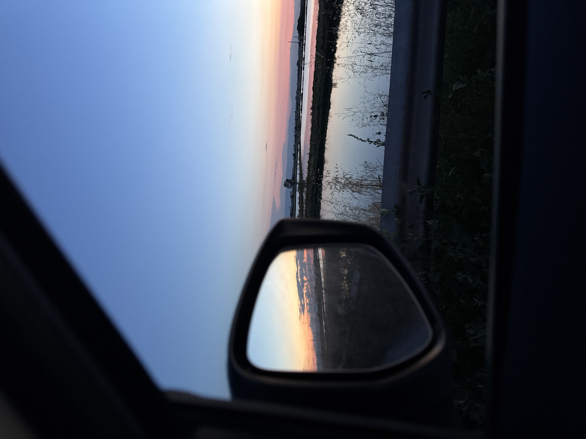 The image is a landscape shot taken from inside a vehicle, looking out the window and into a side mirror. The sky is a gradient of blue, transitioning to pink and orange near the horizon, indicating either sunrise or sunset. In the foreground, the dark frame of the car's interior and the side mirror are visible. The side mirror reflects a portion of the same landscape, showing the sky, clouds, and a body of water.
The wider view shows a body of water, possibly a canal or marshland, with grassy banks. The water reflects the colorful sky. Tall grasses and reeds are visible along the edge of the water. In the distance, there are low-lying hills and some structures, possibly industrial buildings, are faintly visible on the horizon. A metal guardrail runs horizontally across the lower part of the image, separating the foreground from the landscape. The overall impression is one of a serene, natural scene viewed during twilight. There are no people or animals visible in the image.