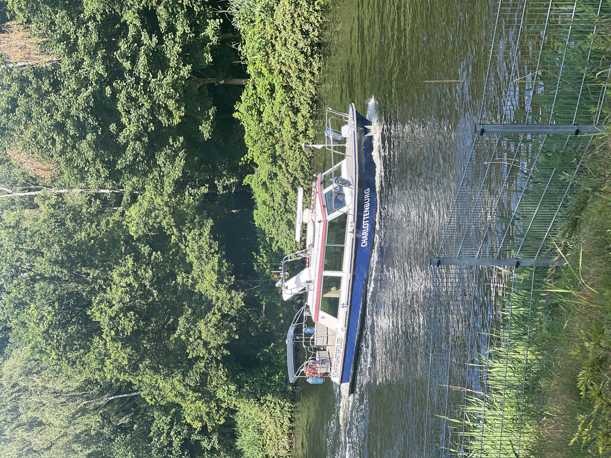A police boat, marked "SCHIFFAHRTSPOLIZEI CHARLOTTENBURG," travels down a waterway, creating a wake. The boat is white with blue and red trim, and has a covered seating area at the stern. It is equipped with navigational equipment. The waterway is bordered by lush green trees and vegetation on both sides. In the foreground, a metal fence with a wire mesh runs along the grassy bank. The sky is clear and the sun is shining. The scene appears to be in a natural setting within or on the outskirts of a city.
