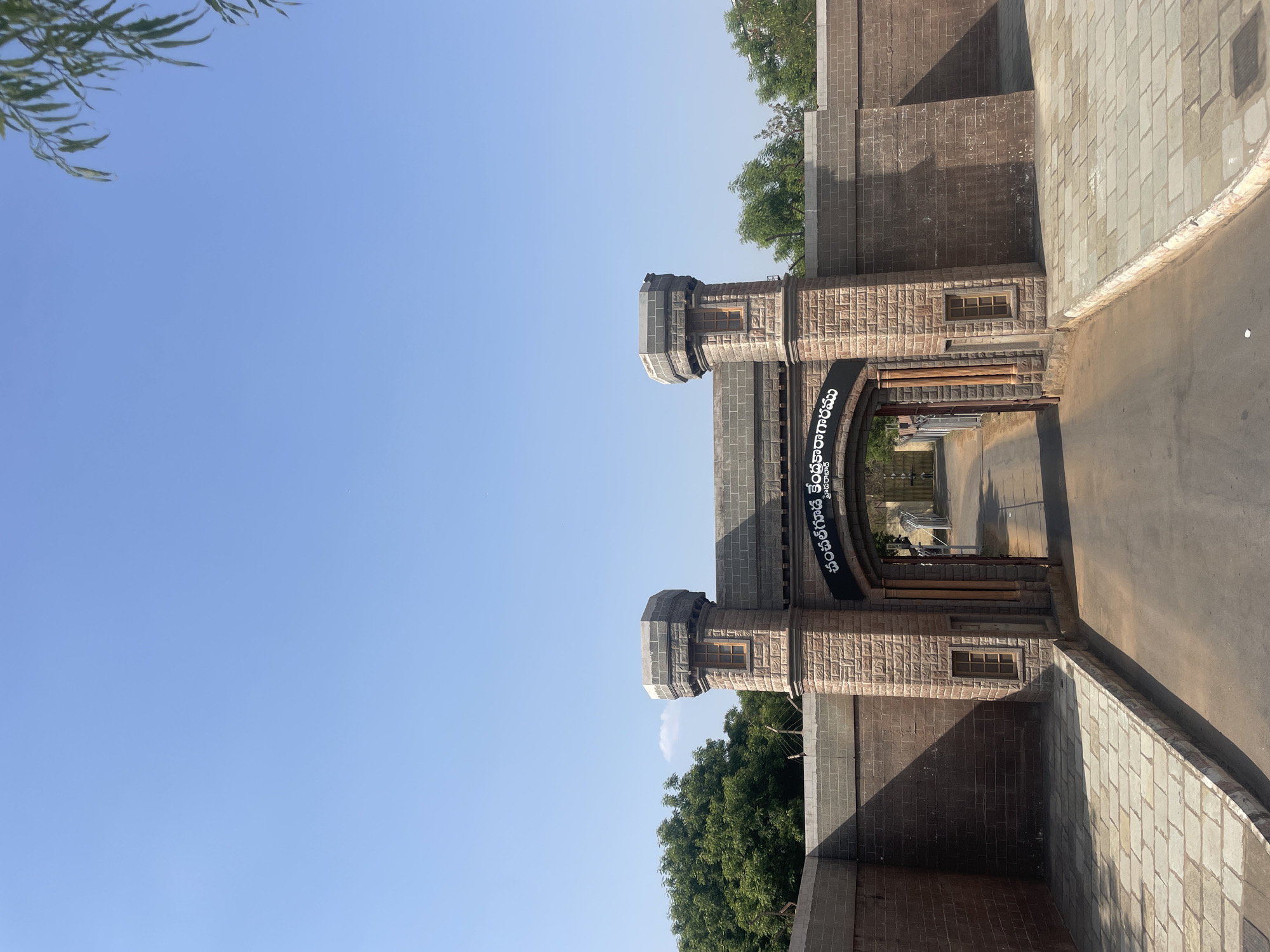 A low-angle, horizontally-oriented photograph depicts the entrance gate to the District Central Jail in Anajpur, India. The gate is constructed of tan stone or brick with darker gray accents, featuring two towers flanking a central archway. A sign, likely in Telugu script, spans the arch. Beyond the gate, a road or walkway extends into the prison complex. The sky above is clear and blue, with foliage visible at the top left and behind the gate structure. A paved area is on the right side of the road.