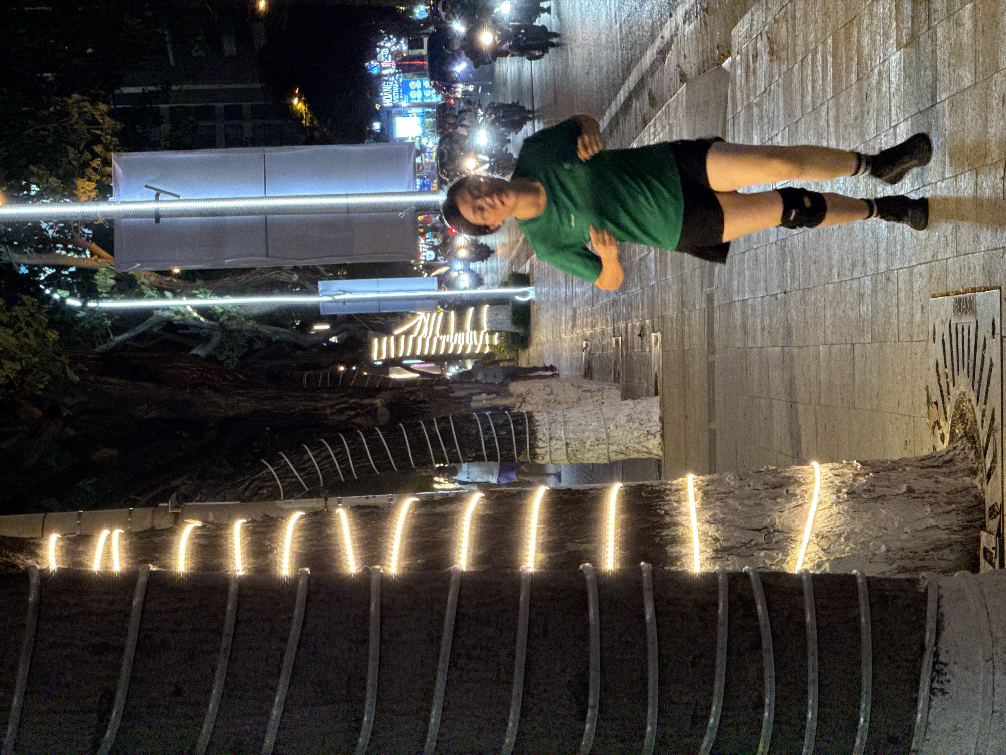 A woman is jogging on a paved sidewalk at night in Nha Trang, Vietnam. The sidewalk is lined with trees decorated with string lights. The woman is wearing a green t-shirt, black shorts, a knee brace, black socks, and black athletic shoes. She is looking forward and appears to be mid-stride. In the background, there are motorcycles with headlights on, and buildings with illuminated signs, indicating an urban setting. The street is wet, suggesting recent rain.