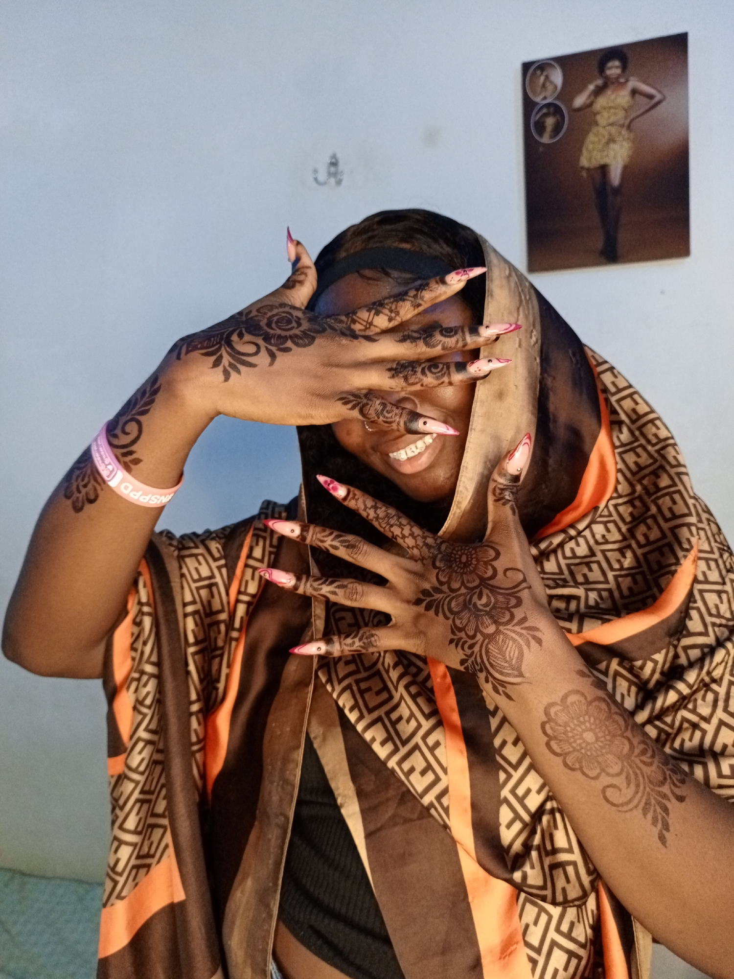 A medium shot photograph shows a woman with dark skin partially obscuring her face with her hands, which are decorated with intricate henna tattoos. She wears a patterned headscarf and a pink wristband on her left arm. Her long, sculpted fingernails have a pink design. She is smiling slightly, and her teeth are visible through the gaps between her fingers. The background is a plain white wall with a small framed picture of a woman in a dress hanging on it. The location is Unknown, Unknown.