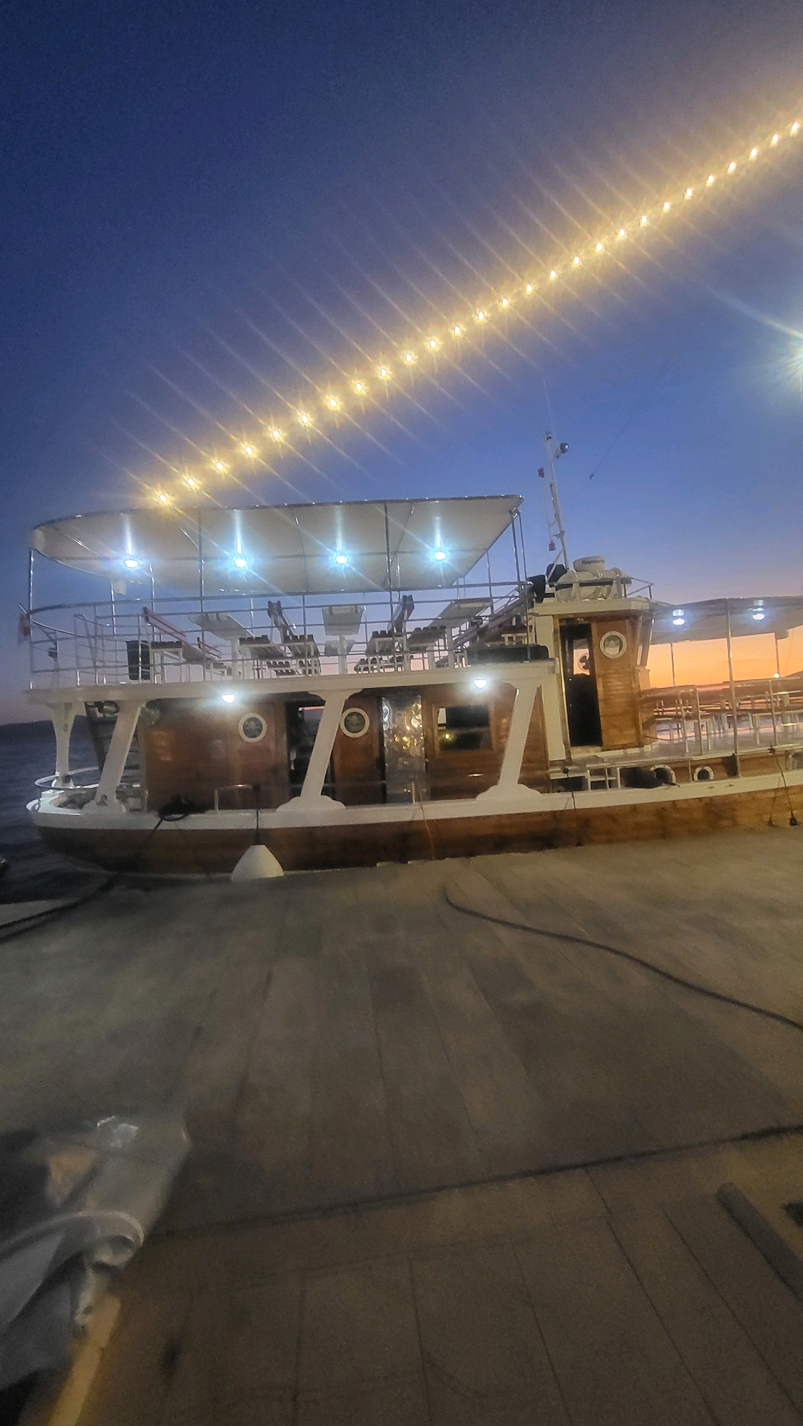 A wooden boat with a white canopy is docked at a pier at dusk. String lights are visible in the dark blue sky above. The boat has multiple decks with tables and chairs. The pier is made of wooden planks. The water is calm and reflects the sky. The location is Općina Tučepi, Croatia.