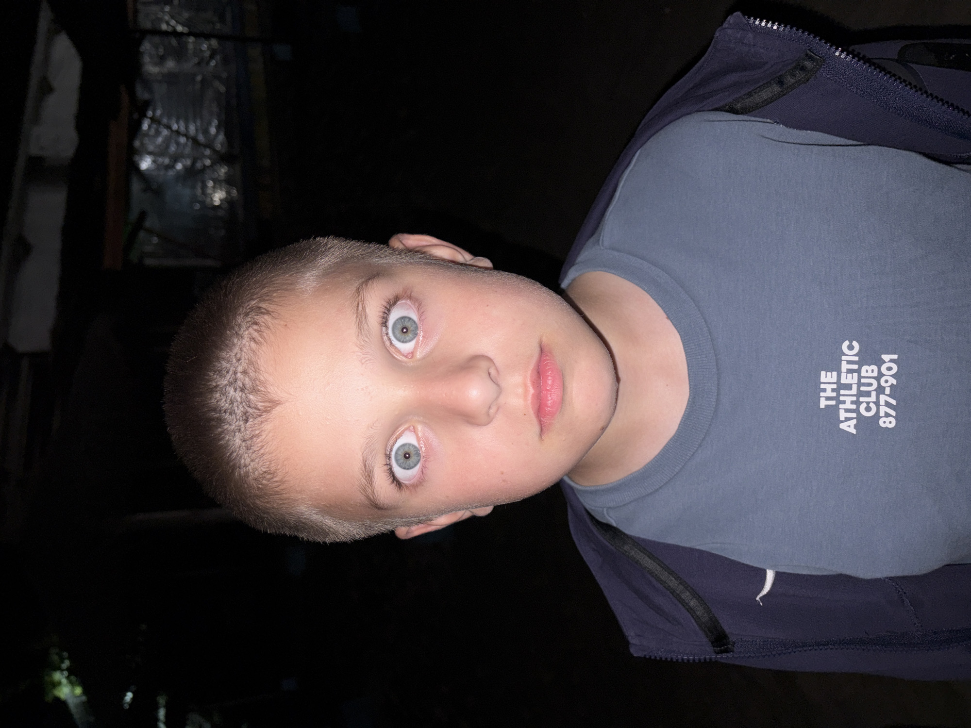 A young male individual, appearing to be a child, is centered in a close-up, chest-up portrait against a dark, indistinct background in Tsarevo, Bulgaria. The subject has short, light-colored hair and fair skin, illuminated by direct frontal flash photography. His light blue-grey eyes are wide open, pupils dilated, staring directly forward. His mouth is slightly parted. He is wearing a dark blue zippered hooded jacket over a light blue crew-neck t-shirt. The t-shirt bears white text, reading "THE ATHLETIC CLUB" above "877-901". The background is predominantly black, with faint, out-of-focus, dark structures and sparse, reflective elements visible in the upper and right portions, indicating a dimly lit outdoor or semi-enclosed environment at night.