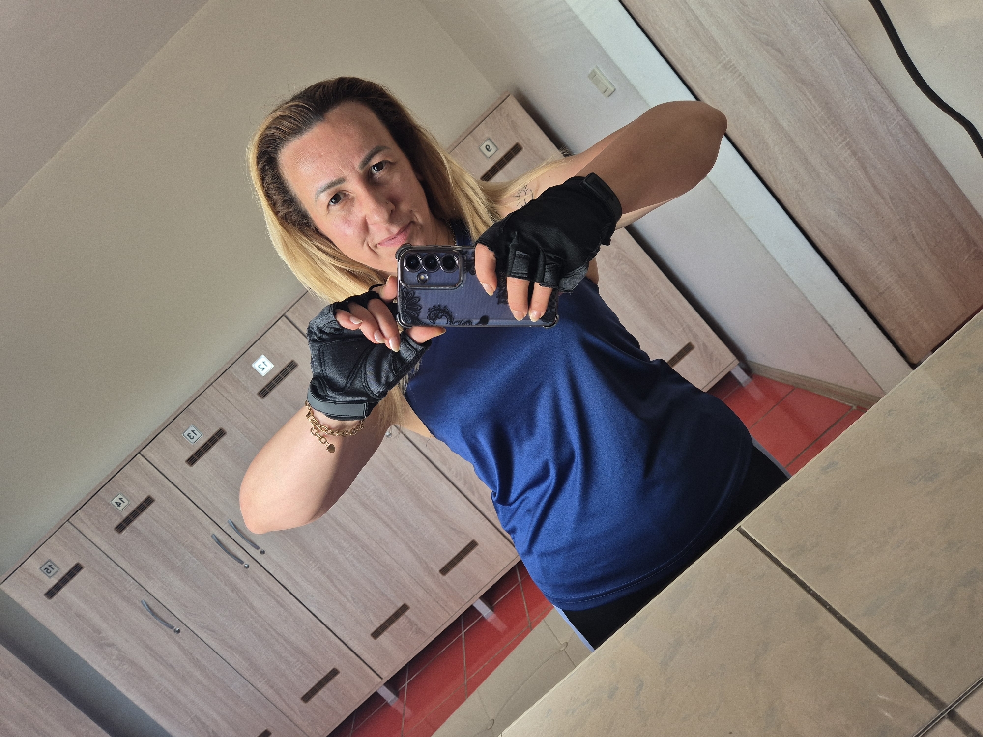 A woman with blonde hair and fair skin takes a selfie in a locker room. She is wearing a blue tank top and black fingerless gloves, and she has a gold bracelet with a heart charm on her left wrist. She is holding a smartphone with a floral case in her right hand. Behind her are wooden lockers, and the floor is red. The image is taken from a low angle.