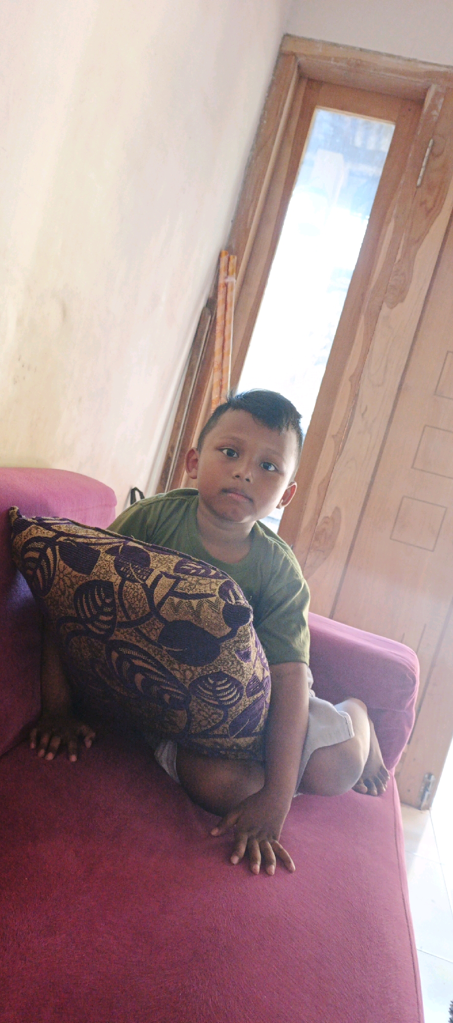 A young boy sits on a vibrant red sofa, leaning against a patterned pillow. He is wearing a green t-shirt and light-colored shorts. His hands are resting on the sofa, with his fingers splayed. The background shows a wall and a wooden door frame with a window. The scene appears to be indoors, likely in a residential setting within Kasiyan, Indonesia, given the distinctive wooden door and interior style. The lighting suggests it is daytime, with natural light entering from the window.