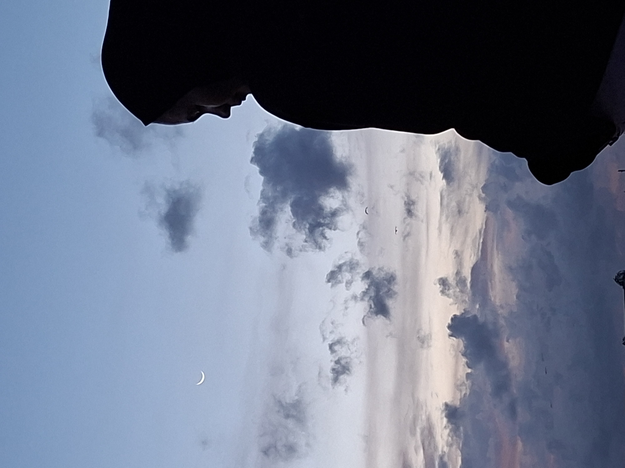 The image, taken in what appears to be Lhokseumawe, Indonesia, shows a silhouette of a person wearing a hood against a background of a partly cloudy sky. A crescent moon is visible in the lower left corner. The clouds vary in color from light grey to dark grey. There are also some dark birds flying in the sky. The image is tilted to the left. The image's orientation suggests it was captured looking upward towards the sky.