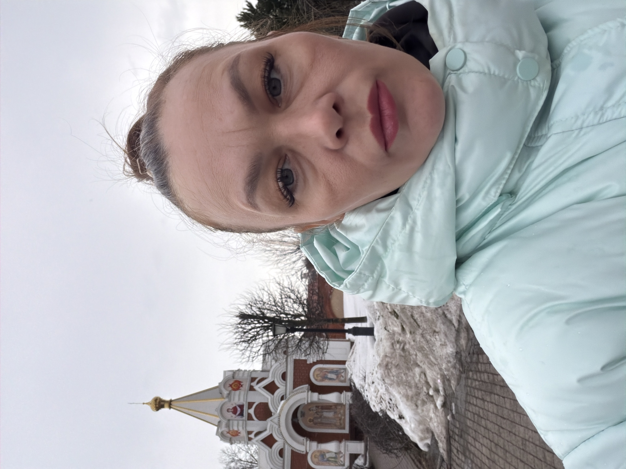 An adult female is depicted in a selfie, positioned in the foreground. She appears to be in her 30s or 40s, with medium skin tone, brown hair styled in a bun with visible grey strands, and is wearing extended upper eyelashes and reddish lipstick. She is dressed in a light mint green puffer jacket, which exhibits visible water droplets, over a dark underlayer. Her expression is neutral.
In the mid-ground, the Chapel of the Holy Royal Martyrs in Izhevsk, Russia, is visible. The chapel is a red brick structure with white architectural trim, featuring a single gold-colored dome topped with an Orthodox cross. The facade includes multiple arched niches containing painted religious icons. To the left of the chapel's entrance, a black, ornate street lamp stands.
The ground around the chapel and in the foreground consists of a paved path and irregular patches of dirty, melting snow. Bare deciduous trees are present in the background, to the left and right of the chapel. The sky is overcast.