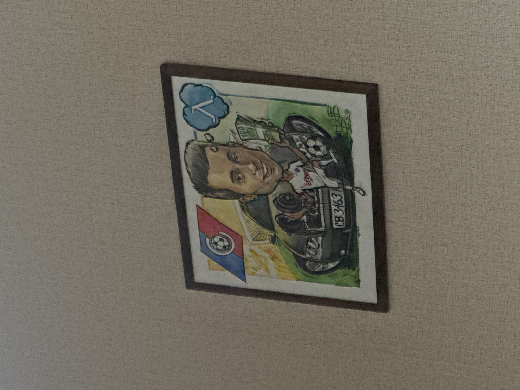 A framed watercolor caricature is hanging on a textured wall. The caricature depicts a man's head and upper body emerging from the front of a black car. The man has short, dark hair and a wide smile. He is wearing a white sports jersey with red trim and appears to be holding a wad of money in his left hand and a soccer ball in his right. The car's license plate reads "CB 3463". Above the man's head, there is a blue thought bubble containing a symbol that resembles the Greek letter lambda (λ) and a cloud. To the left of the man's head, a flag with a soccer ball in the center, split into red and blue sections, is visible. The background of the caricature includes a soccer goal and a hint of a landscape. A signature and the year "2023" are visible at the bottom right of the artwork. The wall appears to be a light brown or beige color with a subtle, repeating pattern. This image was taken in Sofia, Bulgaria.