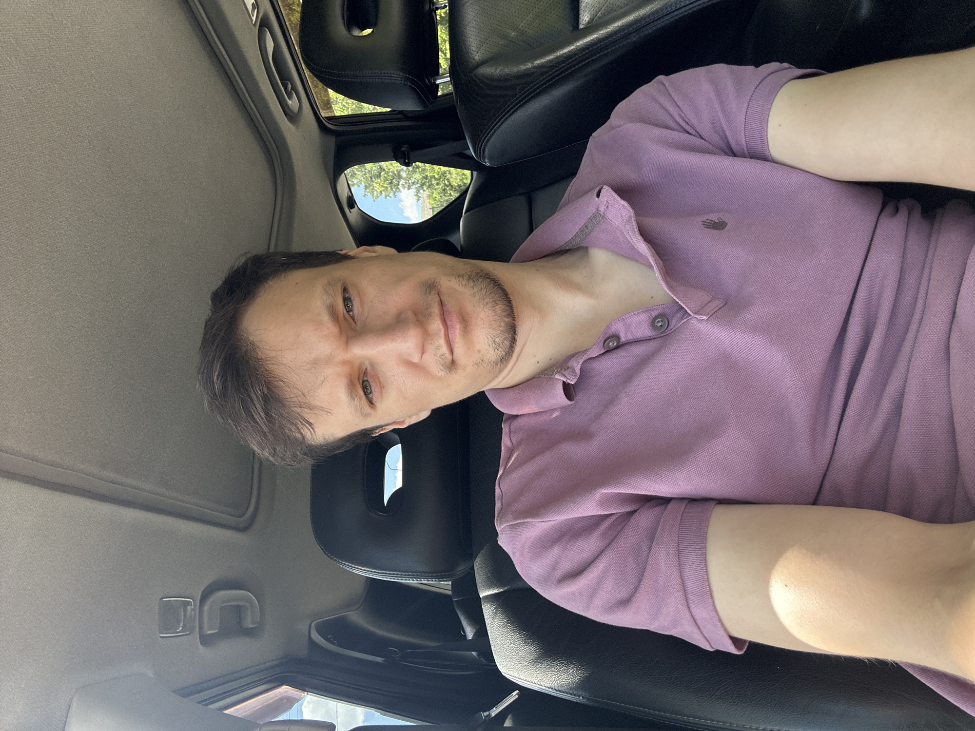 The image shows a man sitting in the driver's seat of a car. He is wearing a purple polo shirt and looking at the camera with a slight frown. The car's interior is visible, including black leather seats, a sunroof, and a handle on the ceiling. Outside the car, trees and a blue sky can be seen through the windows. The scene appears to be a sunny day, and the man is likely in Makiv, Ukraine, based on the provided location.