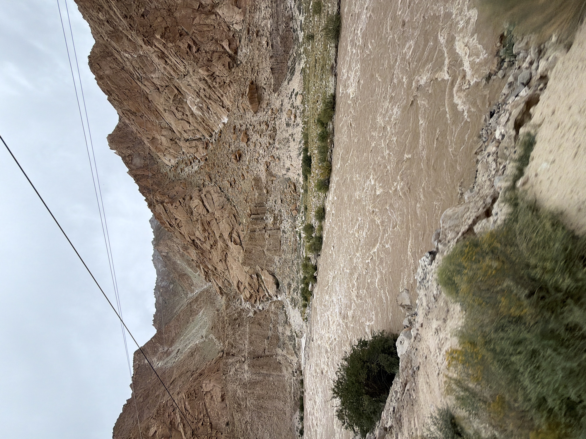 The image depicts a wide, muddy river flowing through a rugged, mountainous canyon under an overcast sky. The river is brown and appears to be moving swiftly, with visible ripples and waves. The canyon walls are steep and rocky, with shades of brown, tan, and gray, indicating a dry, arid environment. Sparse vegetation, consisting of small bushes and grasses, grows along the riverbanks and on the lower slopes of the mountains. A few power lines are visible stretched across the top of the frame. The foreground shows a rocky, uneven embankment with some scrubby bushes. There are no people or animals visible in the scene. The overall impression is one of a natural, untamed landscape in a remote location. The prompt states the location is Hymia, India, which is likely a misspelling or refers to a less common name for a location in the Himalayan region, known for its mountainous terrain and rivers.