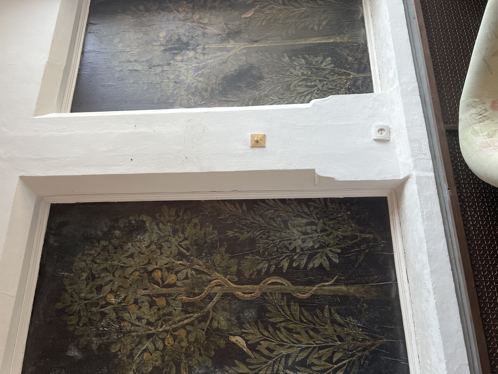 The image shows a section of an interior wall featuring two framed paintings of trees with snakes and birds. The paintings are dark, with green and yellow tones, and appear to be old and somewhat distressed. The wall itself is white and textured, with a white architectural element protruding between the two paintings. On this architectural element, there is a small gold-colored electrical outlet cover and a white circular electrical outlet. The paintings are set into recessed frames. In the foreground, the top of an upholstered chair with a floral pattern is visible. The overall impression is of an older, possibly historic, interior space. The scene is set in the city of Karpaty, Ukraine.