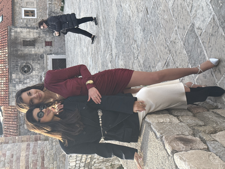 This image captures an outdoor scene within the Old Town of Budva, Montenegro. In the foreground, two adult women are posing. The woman on the left is seated sideways on a low, rough-hewn stone wall. She wears dark sunglasses, a black double-breasted blazer with white accent stitching, a silver-toned chain belt with circular medallions, light beige wide-leg trousers, and black pointed-toe boots. Her left hand rests on the wall, and her right hand is on her thigh. The woman on the right stands beside her, wearing a long-sleeved, V-neck, ruched burgundy mini-dress, a gold wristwatch on her left wrist, rings on both hands, and silver pointed-toe heels with ankle straps. Her left arm is draped over the first woman's right shoulder, and her right hand rests on the first woman's left hip area. Both women look towards the right, away from the camera.
In the mid-ground, a third adult individual walks from the right side of the frame towards the left across a paved stone area. This person wears a dark puffer jacket, dark trousers, and dark athletic shoes, and holds a dark object in their left hand.
The background features historic stone architecture characteristic of Budva's Old Town, including a tall, circular stone tower on the left and a multi-story stone building with a red-tiled roof in the center. This building displays a distinctive circular window (rose window) on its upper level, a rectangular window below, and a ground-level entrance. The ground throughout the scene is paved with irregularly shaped stone slabs. The scene is illuminated by natural daylight, indicating daytime.