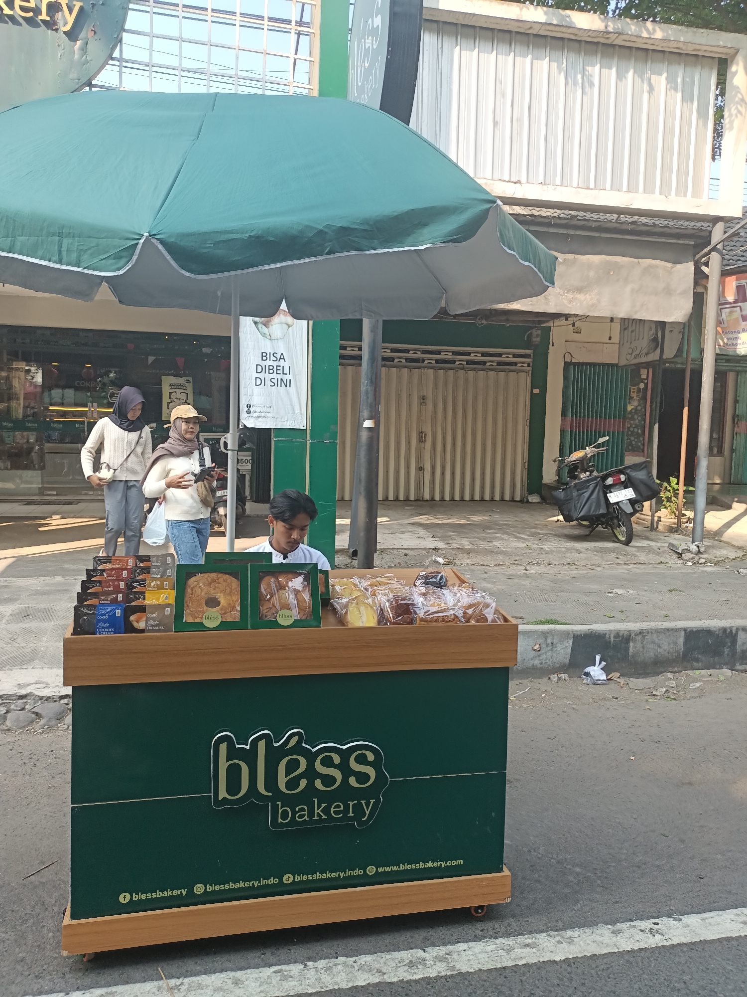 A street-level, medium shot captures a portable bakery cart positioned on a paved roadside in Pare, Indonesia. The cart, painted dark green with a light brown wooden trim, displays various baked goods, including bread and cookies, in clear plastic packaging and green boxes. The cart features the "bless bakery" logo and social media handles.
A young man, dressed in a white collared shirt, stands behind the cart, attending to the products. Two women, both wearing hijabs, are visible in the background. One woman, in a light brown hijab and white sweater, is holding a phone and a bag, and appears to be interacting with the man at the cart. The other woman, in a dark hijab and a cream-colored cardigan, is walking past.
The background reveals a street scene with buildings, including a storefront with a metal roll-up door and a sign that reads "BISSA DIBELI DI SINI" (can be bought here). A motorcycle with black saddlebags is parked on the sidewalk. The environment is illuminated by daylight, casting shadows on the ground. The overall scene depicts a small business operating on a street in a commercial area.