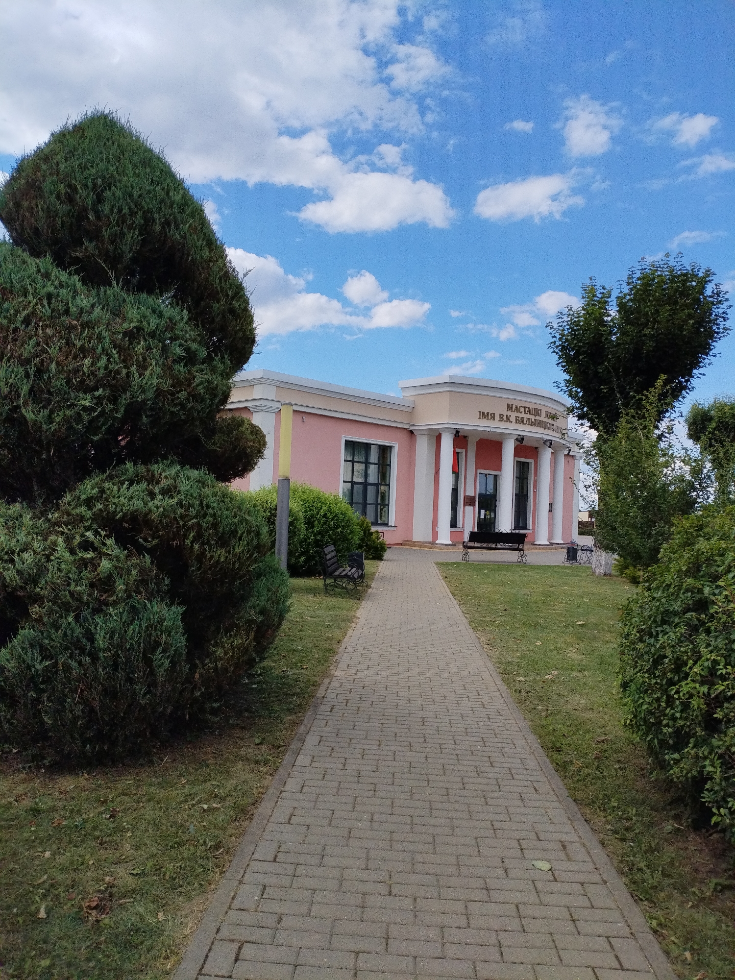 This is a photograph of the Bialynichy City Art Museum, located in Bialynichy, Belarus. The museum is a low-rise building with a pink facade and a white colonnade at the entrance. Above the colonnade, a sign in Cyrillic reads "MUSEUM NAMED AFTER V.K. BALISHCHY". The building is surrounded by a well-maintained lawn and several manicured evergreen bushes, one of which is prominently featured on the left side of the image, sculpted into a unique, rounded shape. A paved walkway leads from the foreground to the museum entrance. A bench is visible on the lawn in front of the museum, and a trash receptacle is next to it. The sky above is blue with scattered white clouds, suggesting fair weather. The overall impression is one of a quiet, cultural institution in a green, park-like setting.