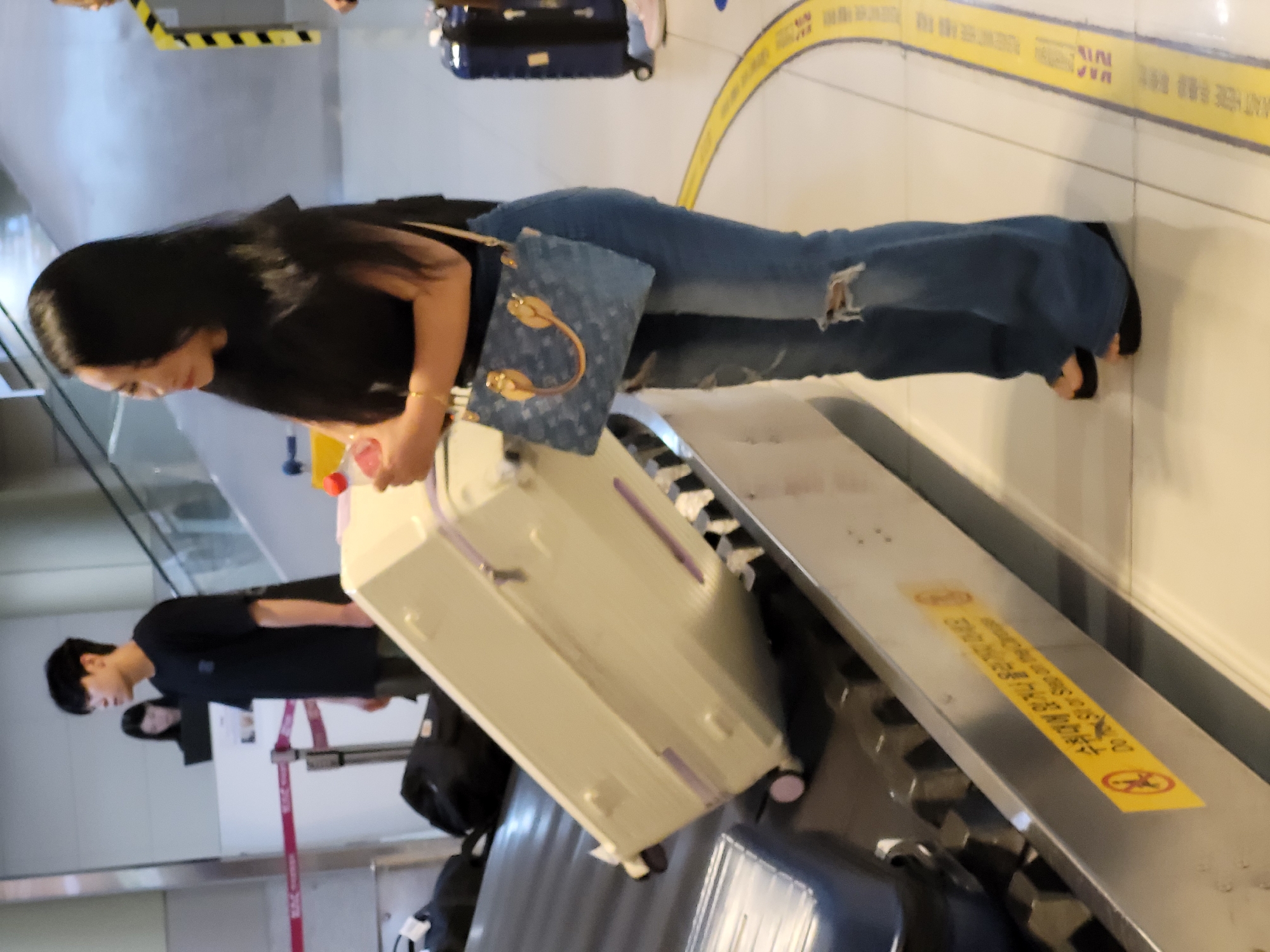 A medium shot shows a person with long black hair wearing a black top and ripped blue jeans. They are holding a blue patterned purse with gold hardware and appear to be standing near a baggage claim carousel. To their left, a large, light-colored suitcase is on the carousel. In the background, another person wearing a black t-shirt and dark pants is visible. A yellow sign with Korean text is partially visible on the carousel. The setting appears to be an airport in Seoul, South Korea, likely at a baggage claim area. The lighting suggests it could be daytime.