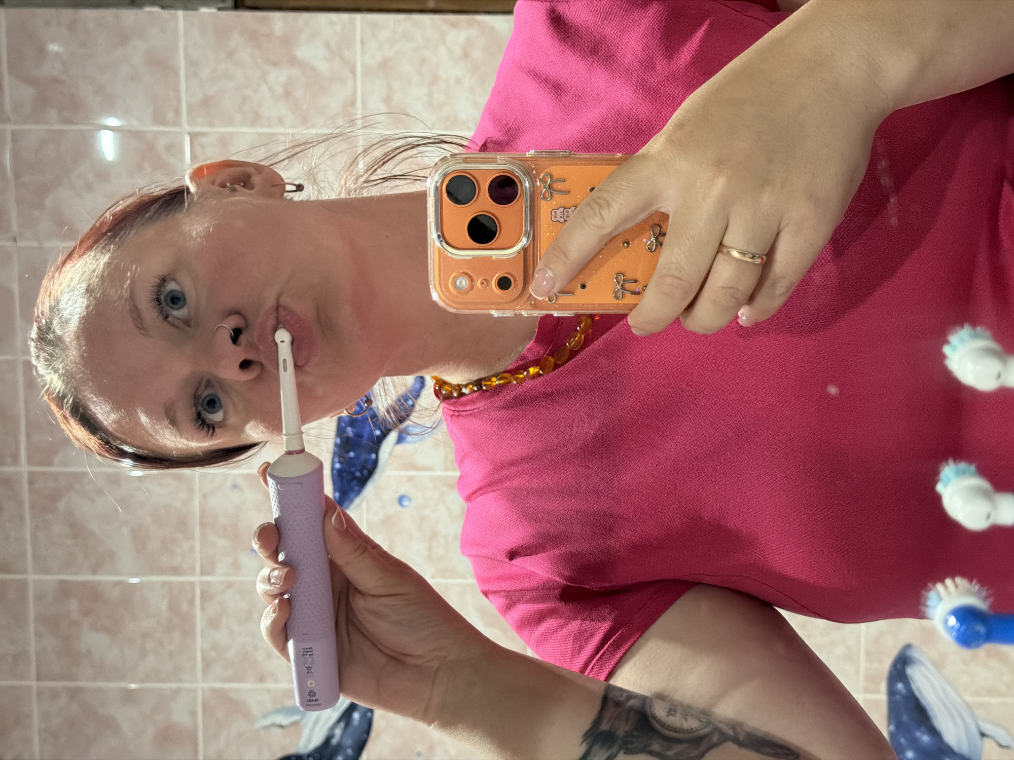 An overhead, mirror selfie captures a person in a bathroom setting, holding a purple electric toothbrush near their mouth. The individual has fair skin, light blue eyes, and reddish-brown hair. They are wearing a bright pink t-shirt and an amber-colored beaded necklace is visible around their neck. The person has a nose ring, multiple ear piercings including a small safety pin in their right ear, and a small gold band ring on their right ring finger. Clear nail polish is visible on their fingernails.
In their right hand, they hold an orange smartphone encased in a clear cover adorned with small decorative bows and a cat-like figure, pointing towards the mirror to take the photo. Their left hand holds the purple electric toothbrush, which has a textured handle with faint, unreadable text and symbols (such as a crossed-out trash can icon) at the base. The white brush head is positioned near their lips, suggesting they are either brushing their teeth or paused during the activity.
The background, reflected in the mirror, shows light-colored square tiles on a wall. On the right side of the reflection, two other electric toothbrushes, one white and one blue, are visible, implying they are on a counter or in a holder. A blue decal or artwork featuring a whale is also visible on the wall below these toothbrushes. Part of a dark tattoo on the person's left forearm is visible, depicting what appears to be a compass or clock face. The overall lighting suggests an indoor setting, likely during the daytime or with artificial light.