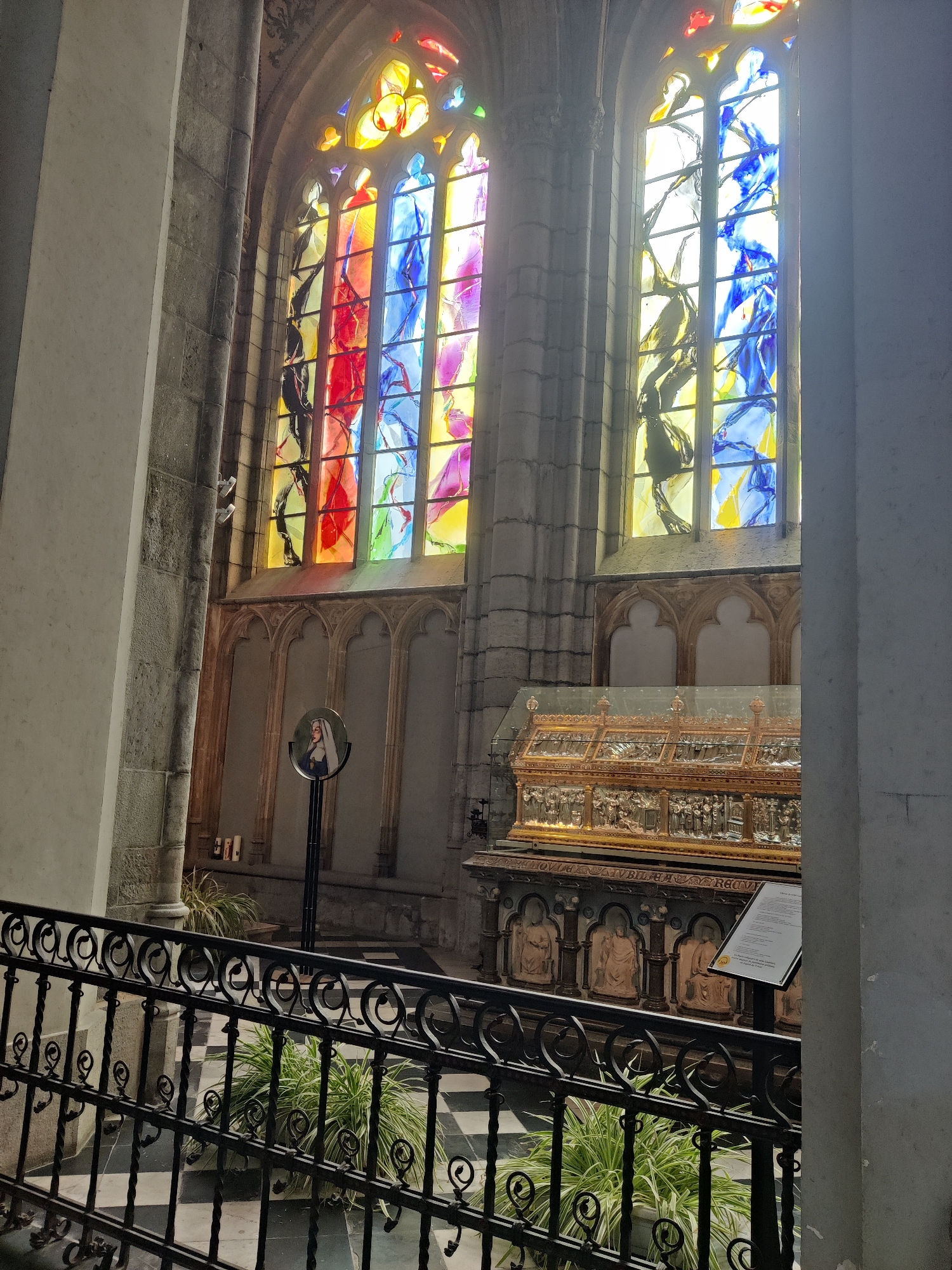 The image shows an interior view of a church in Liège, Belgium, featuring stained glass windows and a reliquary. In the background, two large, arched stained glass windows dominate the upper portion of the scene, illuminated by natural light, casting colorful patterns of red, blue, yellow, and purple onto the stone architecture. Below the windows, a golden reliquary rests on a stone sarcophagus adorned with carved figures and inscriptions. The reliquary is encased in glass and intricately decorated. In the middle ground, a black metal stand holds a circular plaque with an image of a woman in religious attire, likely a nun or saint. A few white candles are visible on a ledge to the left of the plaque. The foreground is occupied by a black wrought-iron railing and a checkered black and white tiled floor. Potted green plants are placed on the floor behind the railing. The overall environment suggests a solemn and historical setting within a religious institution.