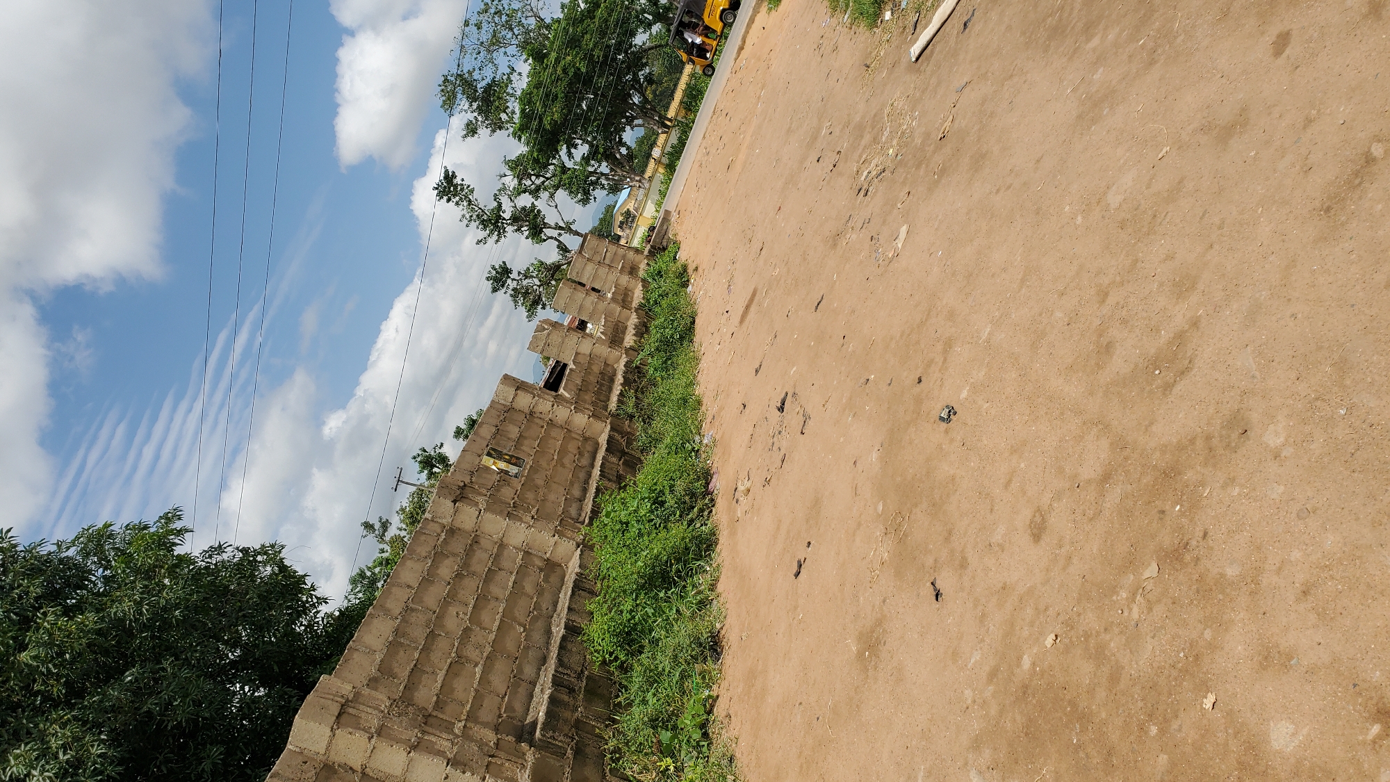 An angled, eye-level shot captures an outdoor scene, likely in Jalingo, Nigeria, under a bright, partly cloudy sky. In the foreground, a dusty, dirt road dominates, scattered with debris and small rocks. To the left, a partially constructed wall made of concrete blocks runs diagonally across the frame, bordered by dense green vegetation. Beyond the wall and vegetation, a paved road is visible, with a yellow three-wheeled vehicle driving on it in the distance. Trees with lush green foliage are scattered throughout the midground and background, and in the far distance, hills are faintly visible. A utility pole is situated behind the construction wall. The overall impression is of a developing or semi-developed area with elements of construction and everyday life.
