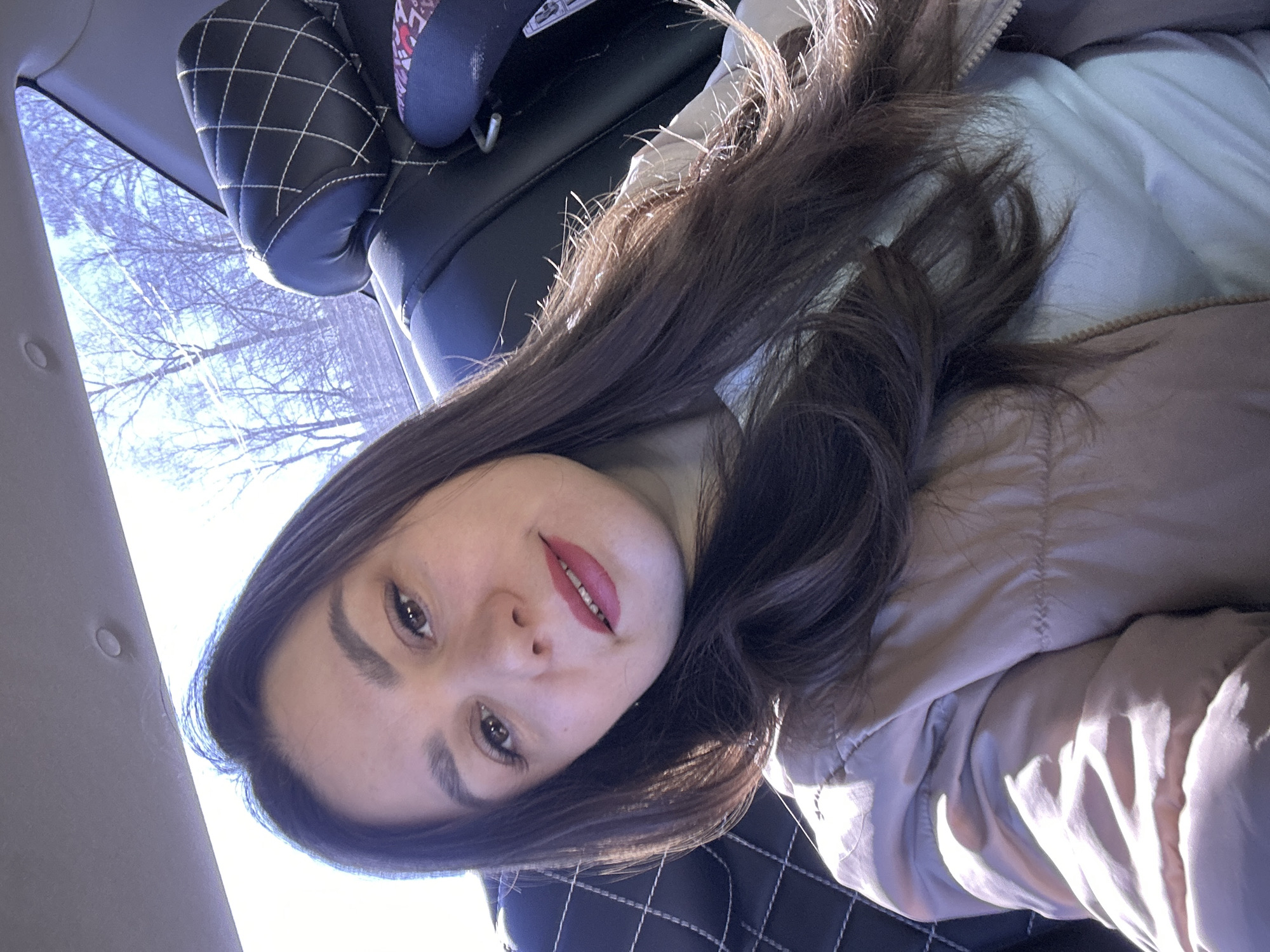 The image shows a woman inside a vehicle. She has dark hair and is wearing a light-colored jacket. The vehicle's interior includes black leather seats with a quilted pattern. The window shows trees outside. The location cannot be determined from the image.