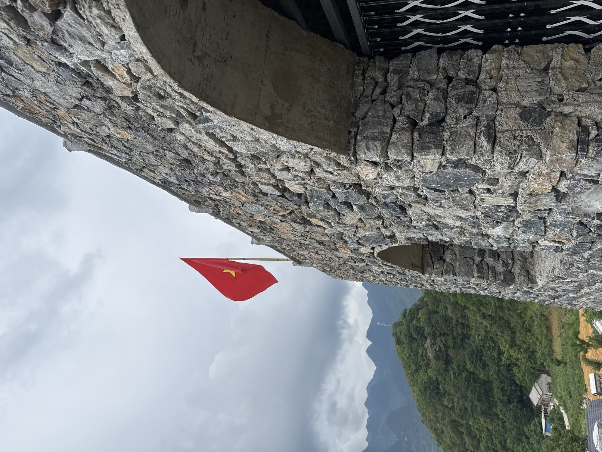 Captured at Lùng Tám Commune, Vietnam, the image presents a low-angle view of a stone structure, likely part of a tower or building. A red Vietnamese flag is prominently displayed, contrasting against the overcast sky. The stonework is composed of irregularly shaped gray stones, indicative of local building materials. The backdrop features a lush, mountainous landscape, characteristic of the region. A glimpse of a building amidst the dense foliage suggests a human presence in the area. The sky is predominantly gray, implying cloudy weather conditions. A metallic structure or railing is visible above the stone arch, indicating a modern addition to the architecture.