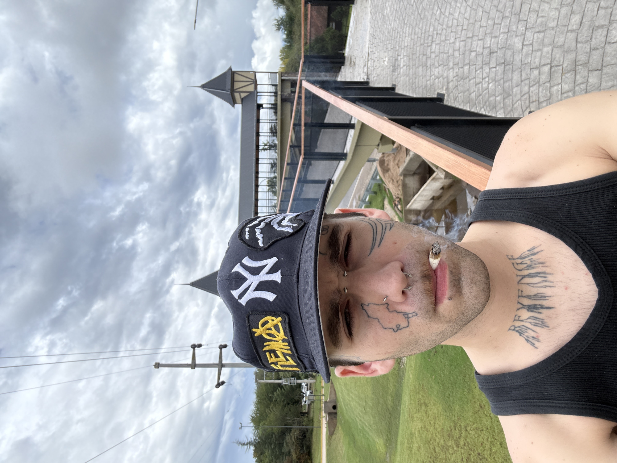 A selfie depicts a man outdoors, likely on a bridge or elevated walkway, smoking a cigarette. He is wearing a black tank top and a black New York Yankees baseball cap. He has multiple facial tattoos and piercings. The background shows a partly cloudy sky, trees, grass, and a building with a distinctive turret. The location is identifiable as Villa General Belgrano, Argentina, based on the architectural style of the building in the background, which resembles traditional German architecture common in that town. The man is holding the phone or camera and taking the selfie. The scene appears to be during daylight hours.