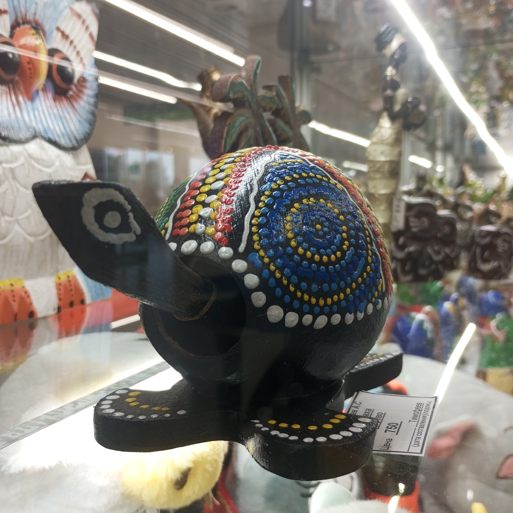 A close-up, eye-level shot shows a painted, wooden turtle figurine with a dark, textured shell decorated with colorful dots and white wave-like patterns. The turtle has black, flat feet adorned with white and yellow dots. A white and blue owl figurine is partially visible to the left, and a tag with Russian text and the number "750" is attached to the turtle's base. The figurine is placed on a glass surface, reflecting a blurred background of shop displays, possibly in a gift shop or market in Naberezhnye Chelny, Russia. The lighting is bright, with a prominent light streak reflecting off the glass.