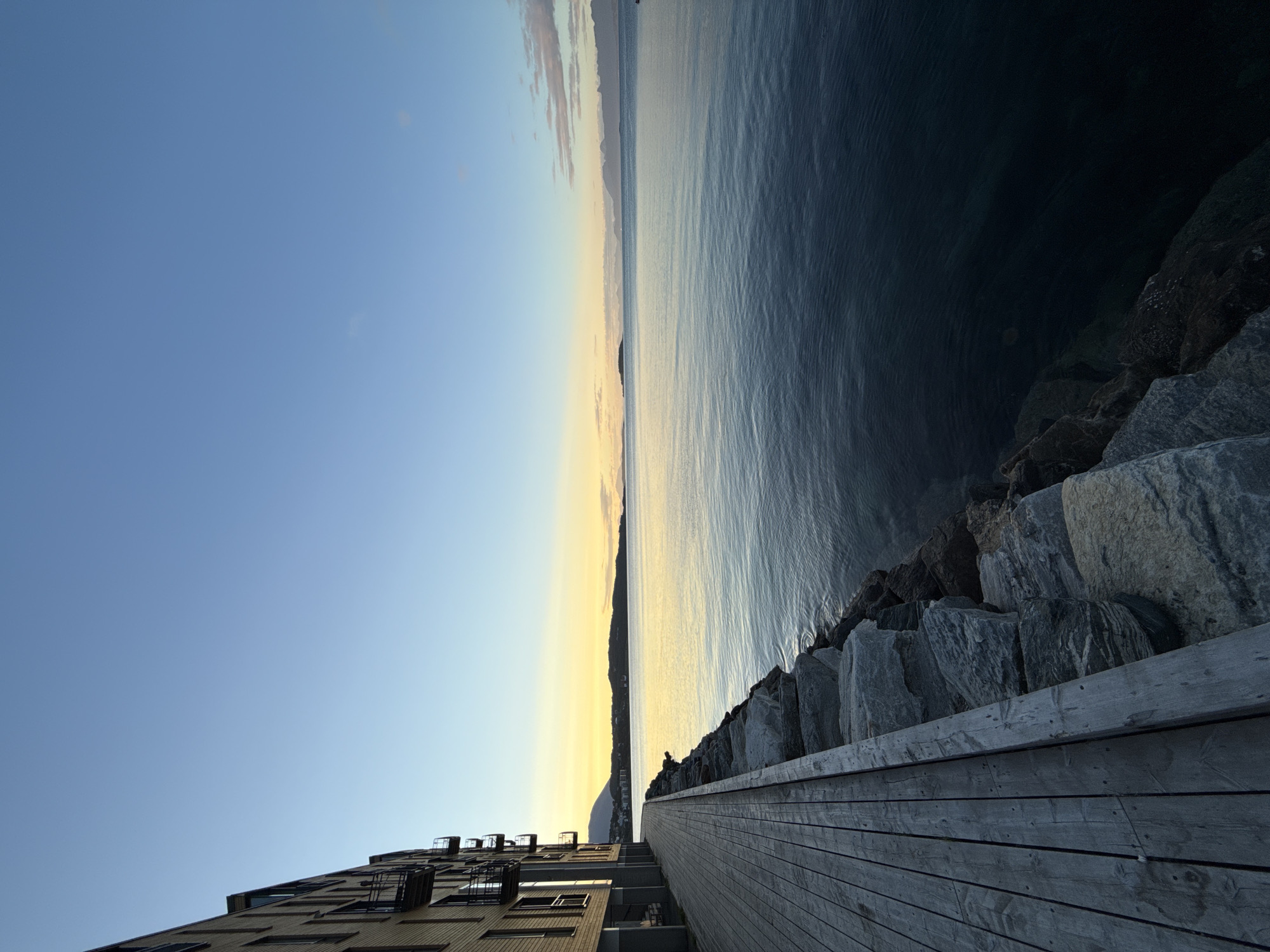 The image shows a wooden boardwalk running alongside a rocky embankment next to a body of water. A modern building with balconies is visible on the left side of the boardwalk. The sky is clear and blue, with a soft yellow glow on the horizon, indicating either sunrise or sunset. In the distance, mountains and islands can be seen across the water. This scene is located in Harstad, Norway, likely along the waterfront promenade known as "Stien langs sjøen" or "The path along the sea." There are no people or animals visible in the image. The environment appears calm and serene.