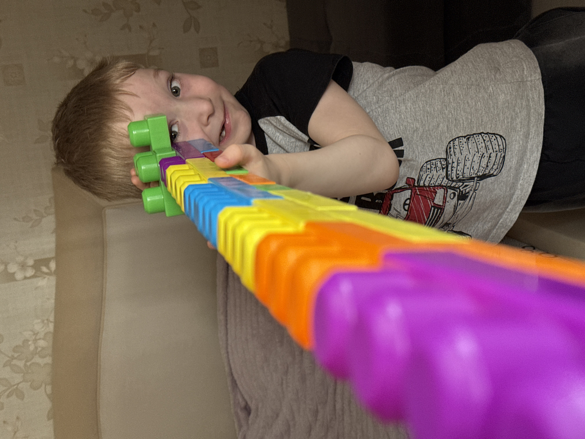 A young, light-skinned child with short blonde hair and green eyes is lying on a beige sofa. The child is holding a colorful plastic toy gun, mostly purple, orange, yellow, blue, and green. The child is wearing a gray t-shirt with a black short sleeve, and black pants. The background includes a beige wall with floral pattern and a dark brown surface behind the child. The location could be a house or apartment in the city of Chaikovsky, Russia.