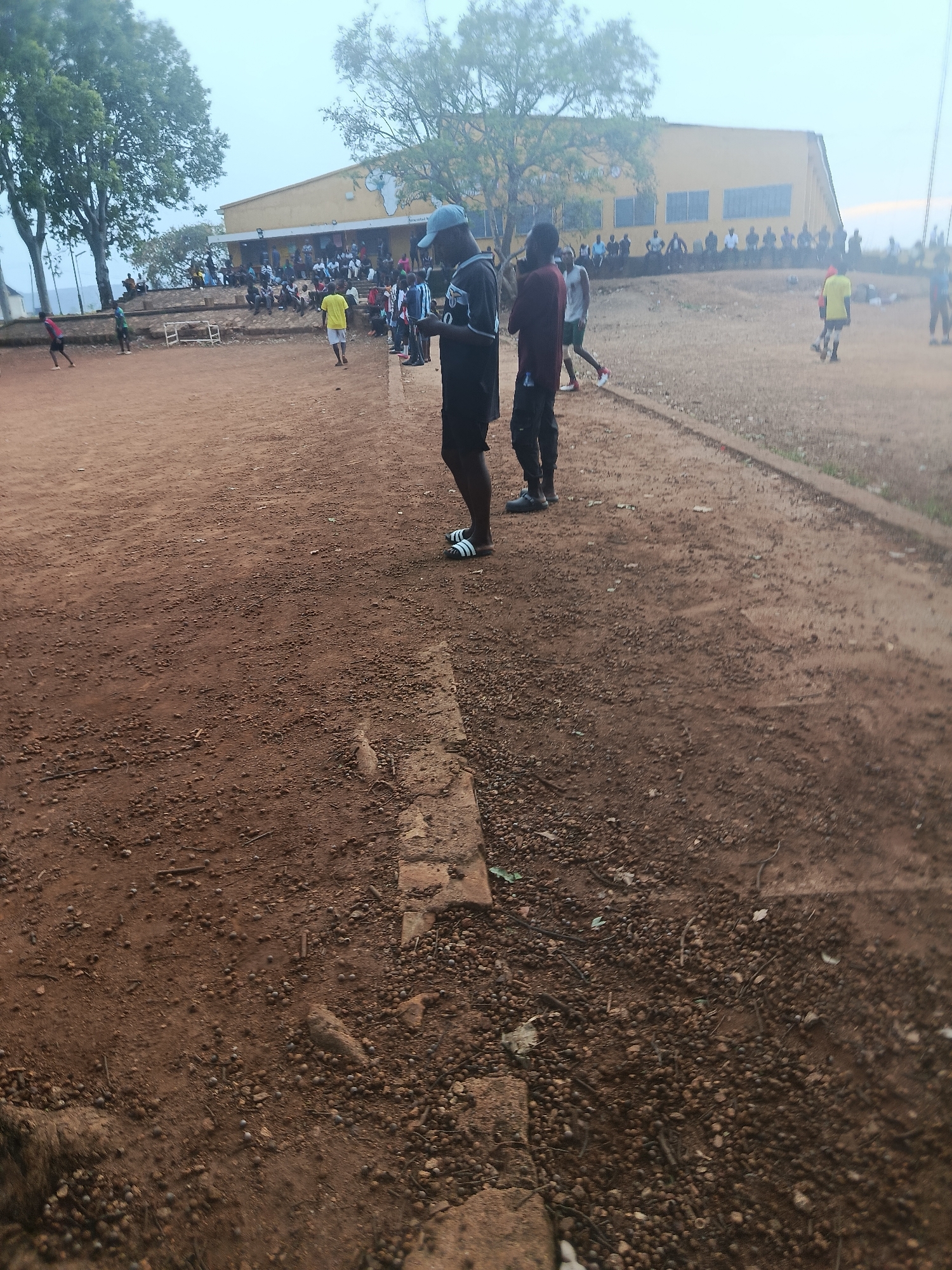 The image depicts an outdoor scene on a dirt field, possibly a football pitch, with a crowd gathered in the background. In the foreground, several individuals are standing and walking on the reddish-brown dirt, which is littered with small pebbles and debris. One person in the immediate foreground is wearing black shorts and a dark t-shirt, with white-striped flip-flops on their feet. To their right, another person is wearing dark pants and a dark t-shirt. Further back, several people are watching from the side of the field. Behind the field is a yellow building with multiple windows and a covered entrance. A line of spectators sits or stands in front of the building. The overall environment appears to be an overcast day with lush green trees visible in the distance, framing the building and the sky. The scene suggests a casual gathering or sporting event. While the location is specified as Rafin Par, Nigeria, no specific landmarks or visual cues within the image directly corroborate this precise geographical identification beyond it being an outdoor setting in what appears to be a somewhat rural or developing area.