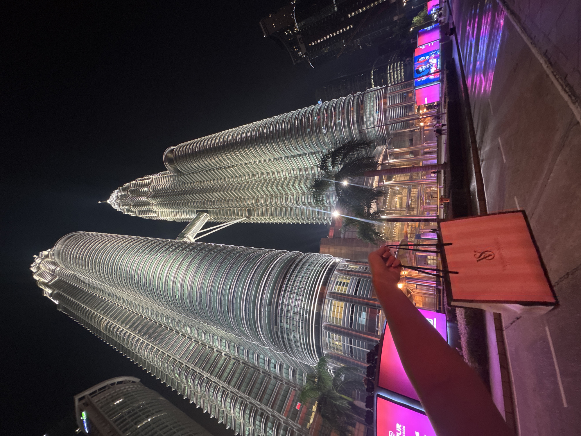 The photograph captures a nighttime scene in Kuala Lumpur, Malaysia, prominently featuring the illuminated Petronas Twin Towers. The towers rise into the dark sky, their intricate facade detail clearly visible. In the foreground, an arm extends upwards, holding a shopping bag with a distinct logo. The ground appears to be a street or walkway, possibly wet, reflecting colorful lights from nearby screens or displays. Other buildings, also illuminated, can be seen in the background. The image suggests a tourist location within the city.