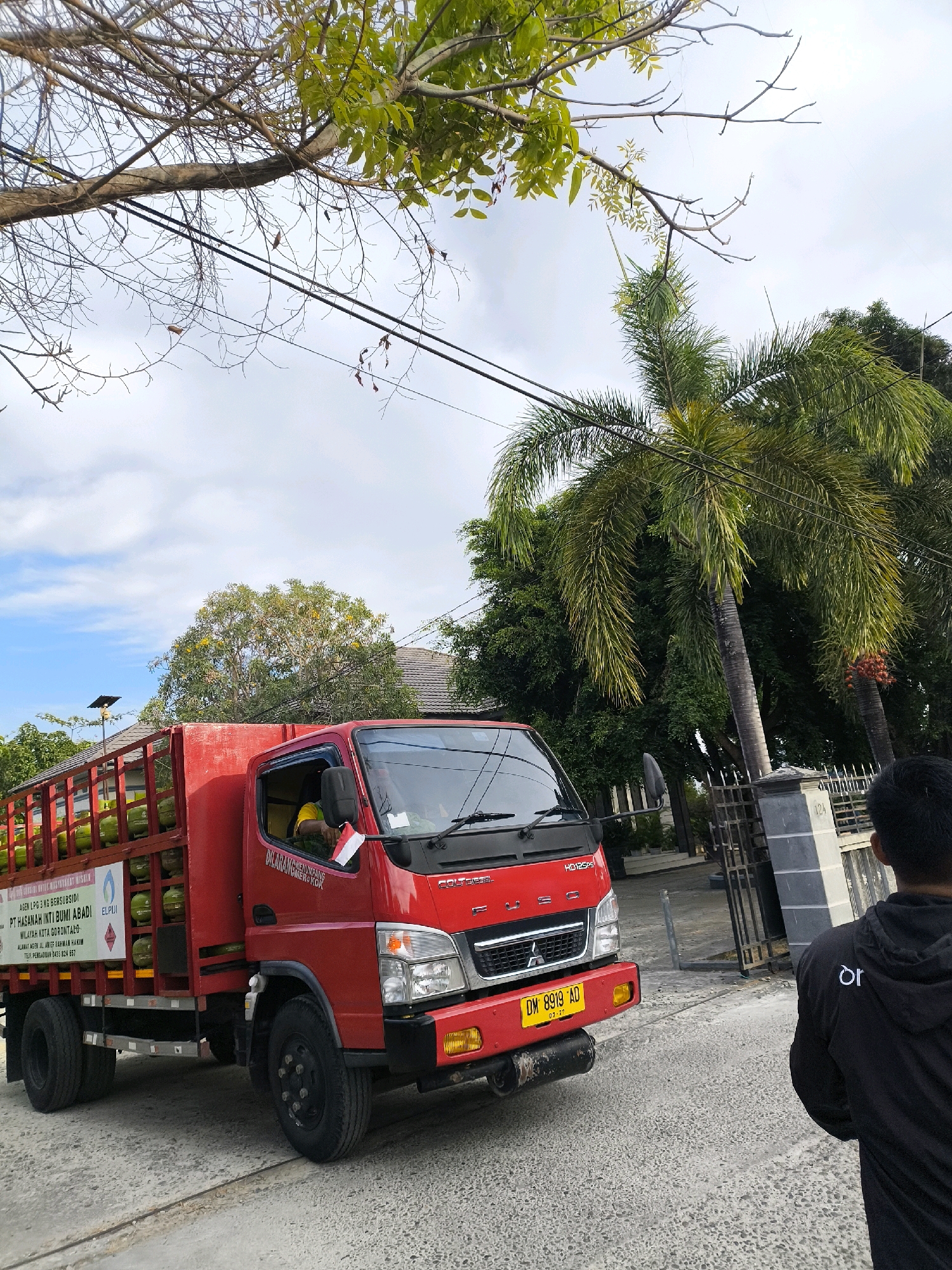 A red Fuso truck is parked on a paved road in what appears to be a residential area in Gorontalo, Indonesia. The truck is loaded with green propane tanks. A man in a black hoodie stands on the right side of the image, facing away from the camera, partially obscuring the truck and a metal gate. The background features houses, trees, and a partly cloudy sky. The truck has a license plate reading "DM 8919 AD" and some text written on its side in Indonesian. A small Indonesian flag is attached to the side of the truck's cabin. The image is taken from a low angle, looking up towards the truck and the surrounding environment.