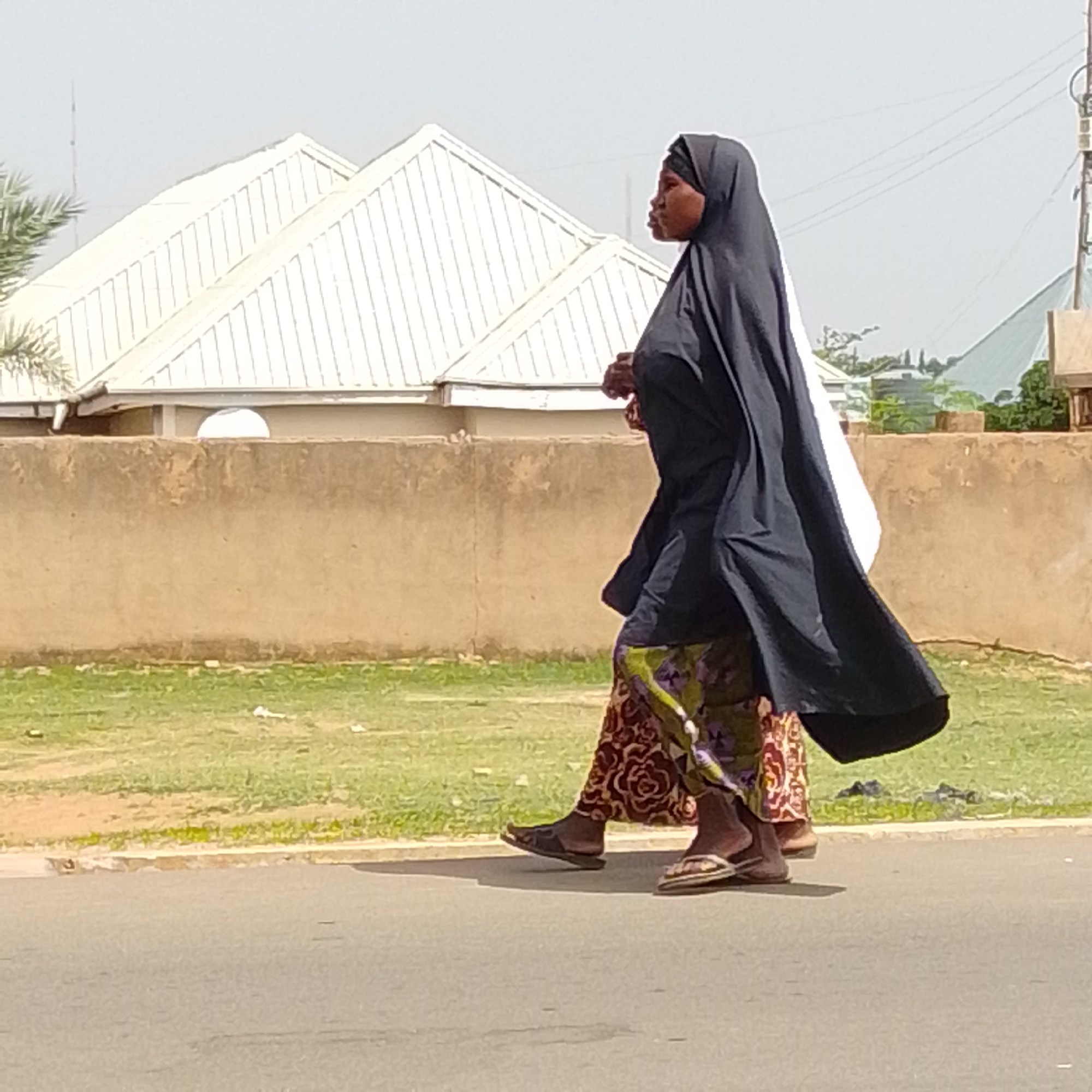 Two women, one in a black hijab and dress and the other in a patterned skirt and a white covering, walk along a paved road in Gombe, Nigeria. A tan-colored concrete wall runs parallel to the road, with a grassy area behind it. In the background, two white, pitched roofs of buildings are visible, along with some scattered trees and power lines against a hazy sky. The scene appears to be a street in a residential or commercial area of the city.