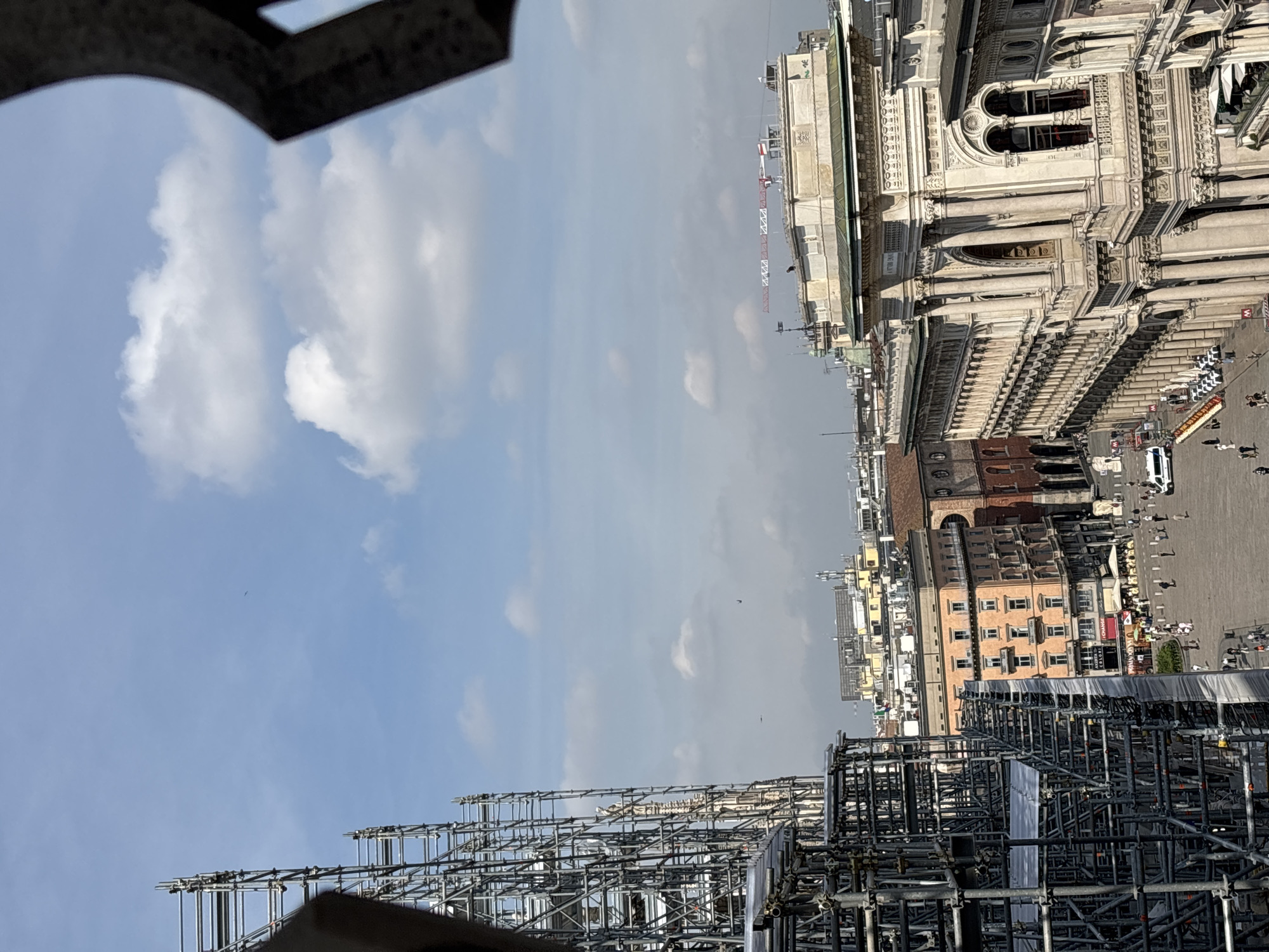 The image shows a view of Milan, Italy, from an elevated position, possibly the roof of the Duomo. The scene includes the Piazza del Duomo, with visible pedestrians and vehicles below. Scaffolding is present in the foreground, obscuring parts of the cityscape. The Duomo itself is visible, showcasing its architecture. The sky is partly cloudy.