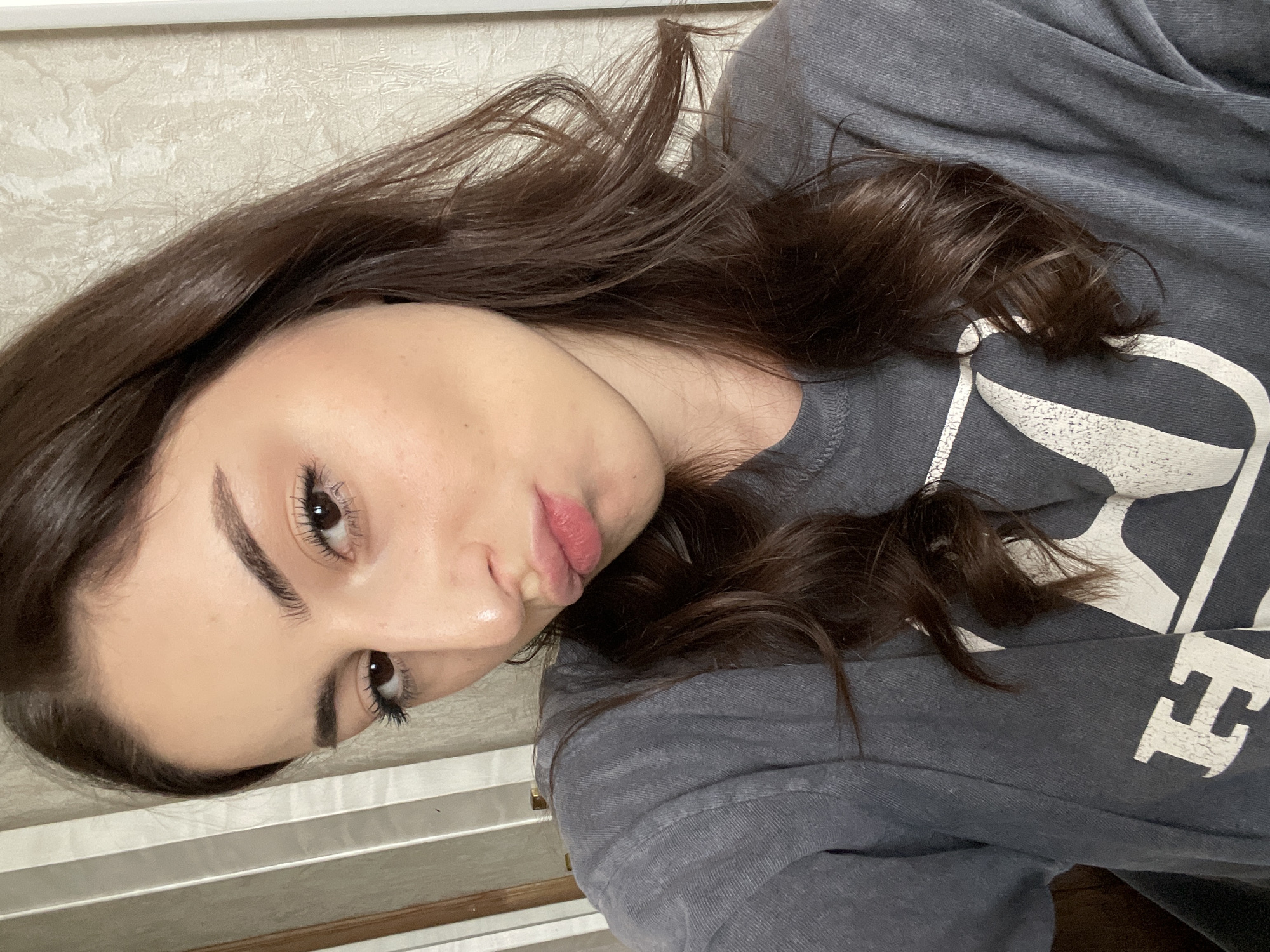 A woman with dark brown hair and brown eyes is lying down and looking at the camera. She has fair skin and full lips. She is wearing a gray shirt with a white design. The background is a white wall with a textured pattern. The image appears to be taken indoors. The location in Moscow, Russia, is unknown.